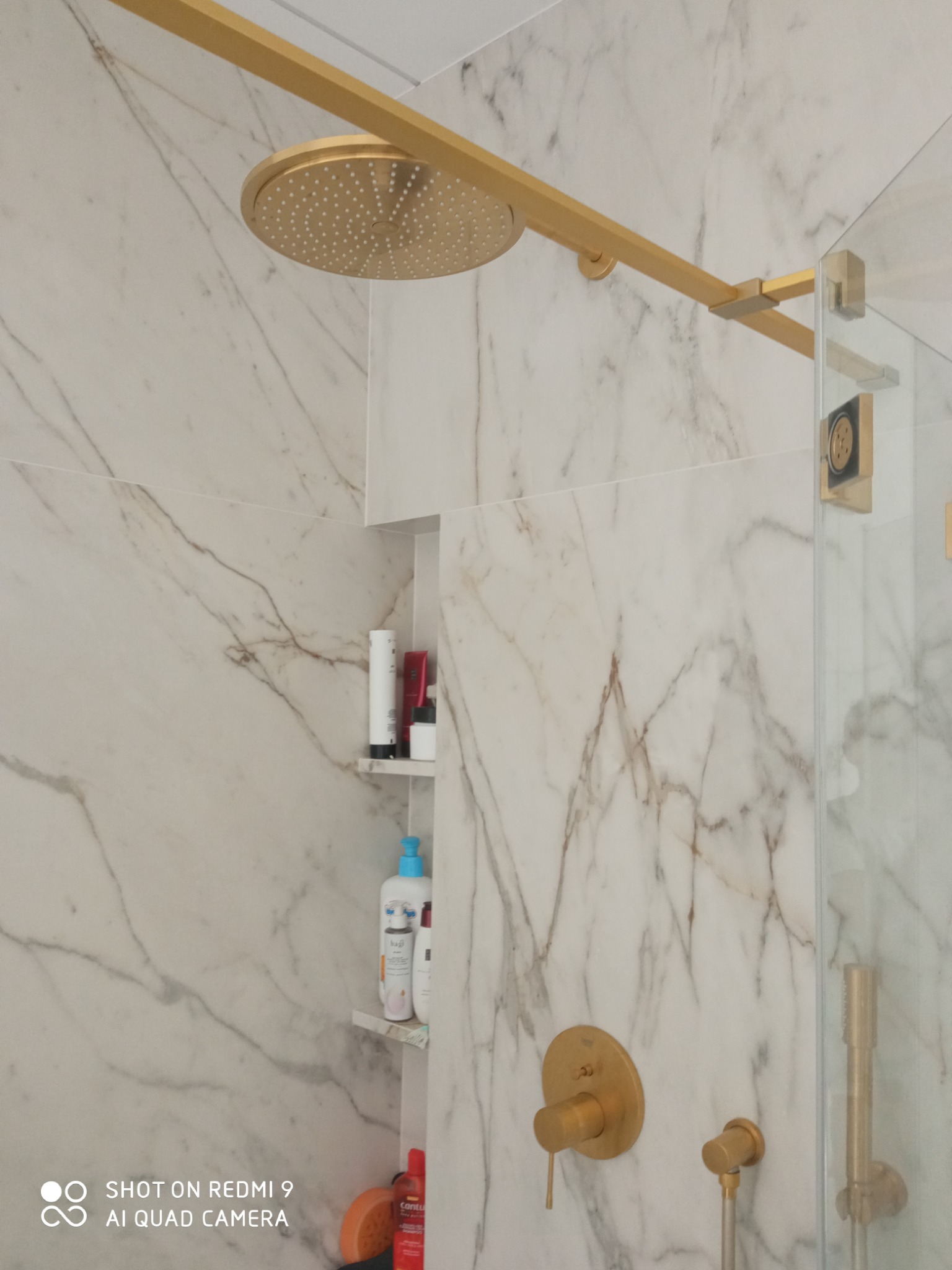 Elegancka kabina prysznicowa z marmurowymi ścianami i złotą armaturą. Widoczne półki z kosmetykami i detale wykończenia.