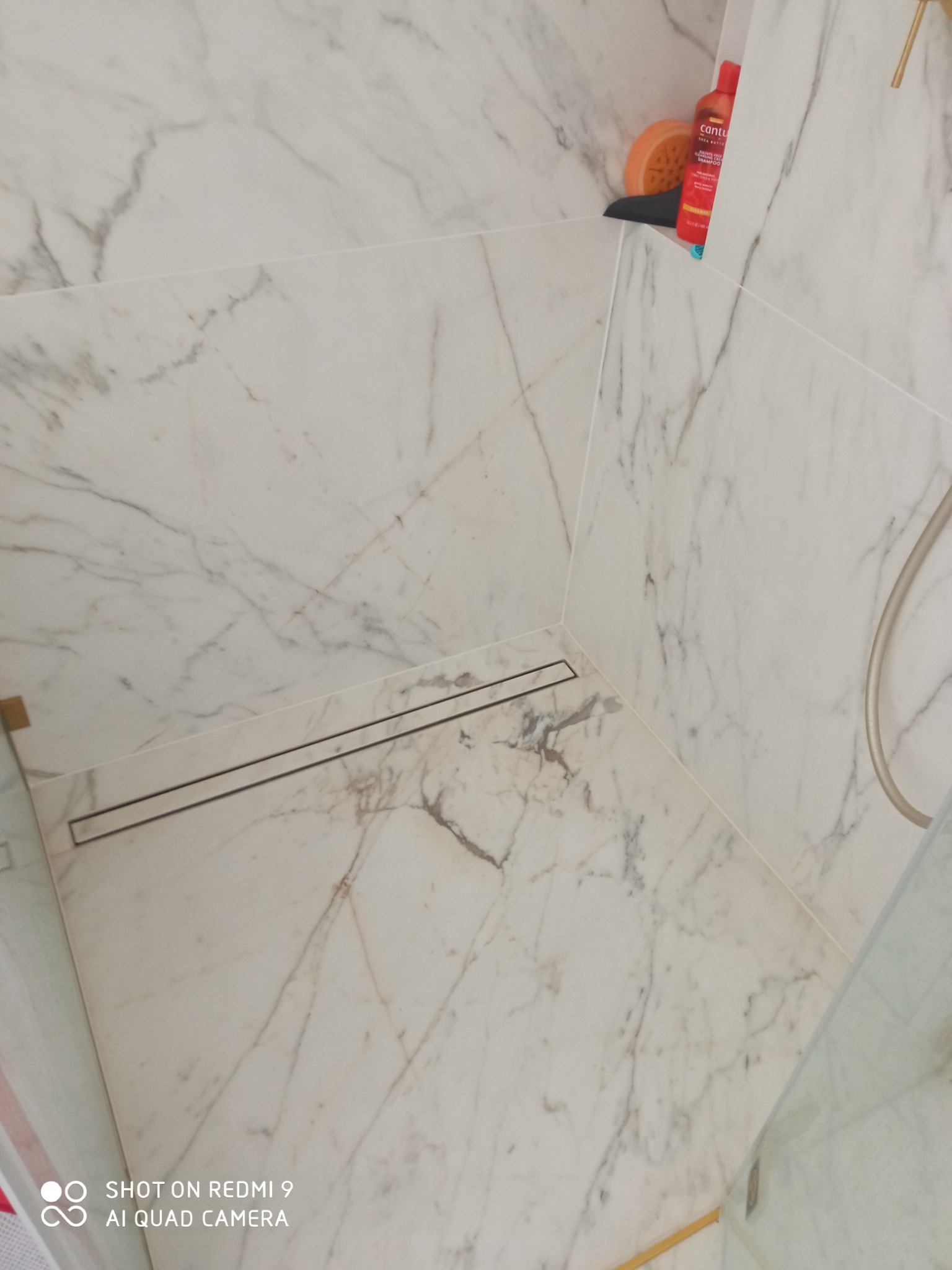 Kabina prysznicowa wykończona białymi płytkami imitującymi marmur, z odpływem liniowym. Widoczny fragment słuchawki prysznicowej i kosmetyki na półce.