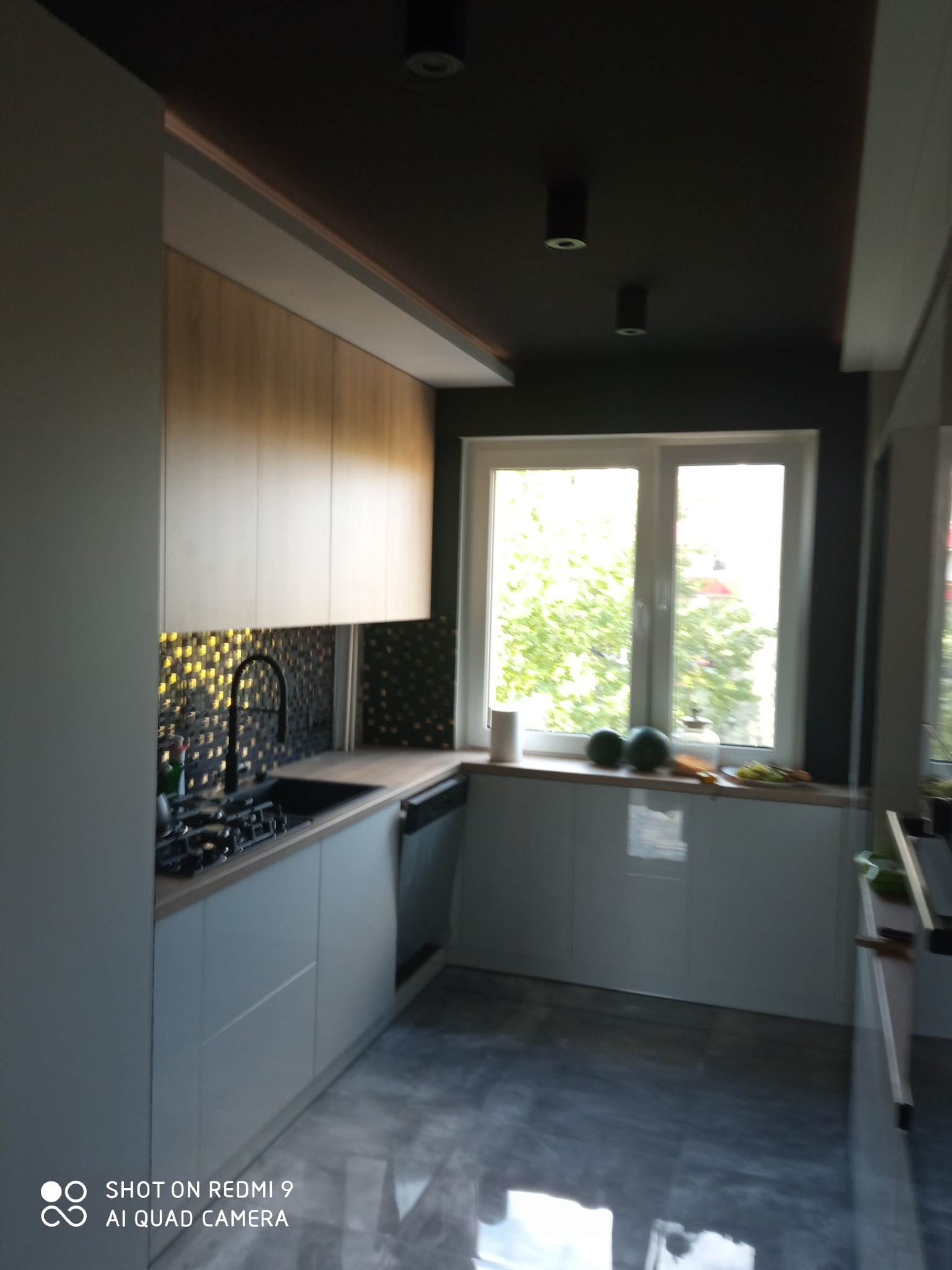 Nowoczesna kuchnia z białymi szafkami, drewnianymi górnymi szafkami i czarną mozaiką nad blatem. Widoczne okno z zielenią na zewnątrz i marmurowa podłoga.