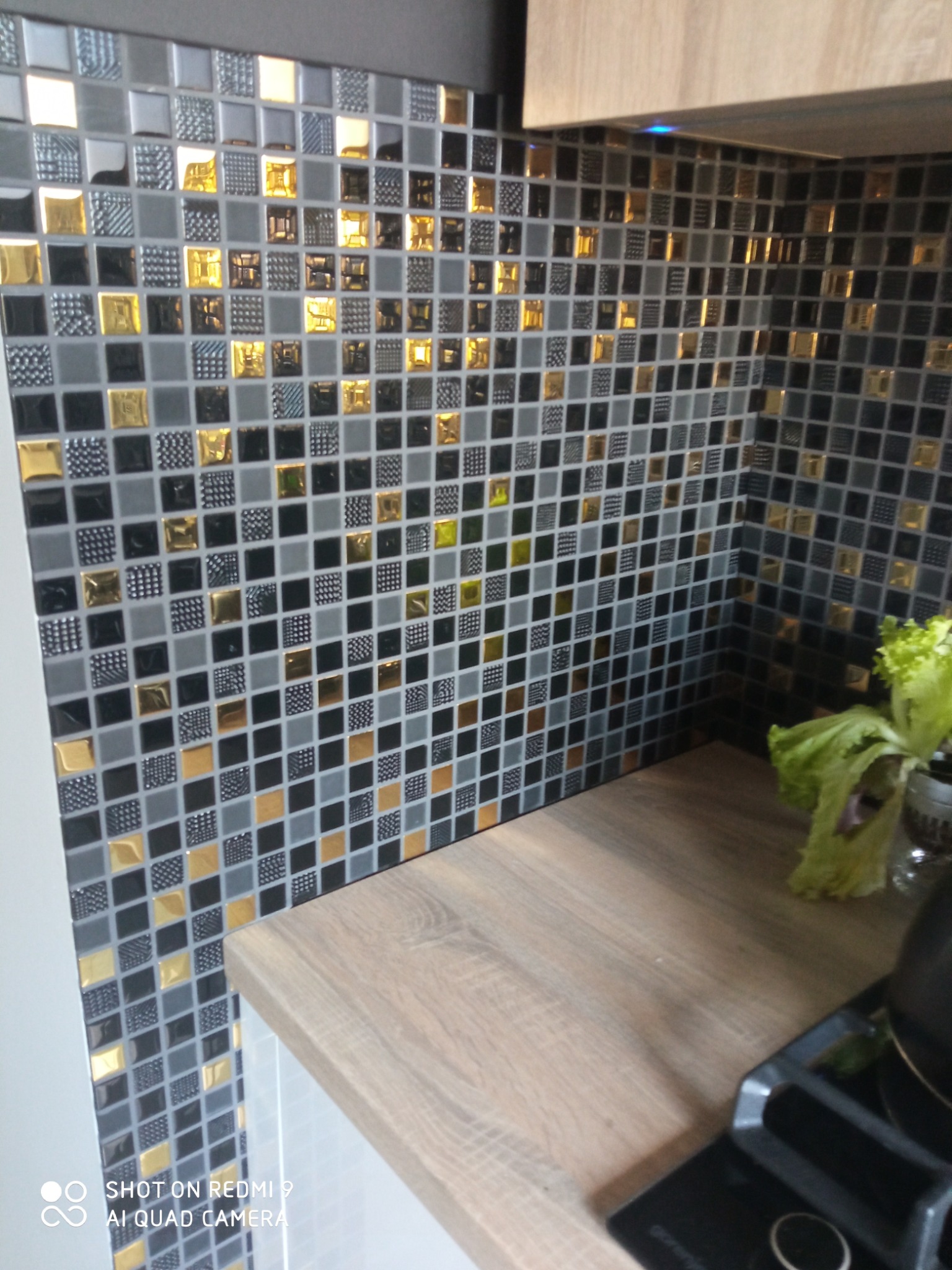 Ściana wyłożona mozaiką w odcieniach szarości, czerni i złota, z blatem kuchennym z jasnego drewna. Widoczny fragment płyty grzewczej i zieleń w tle.