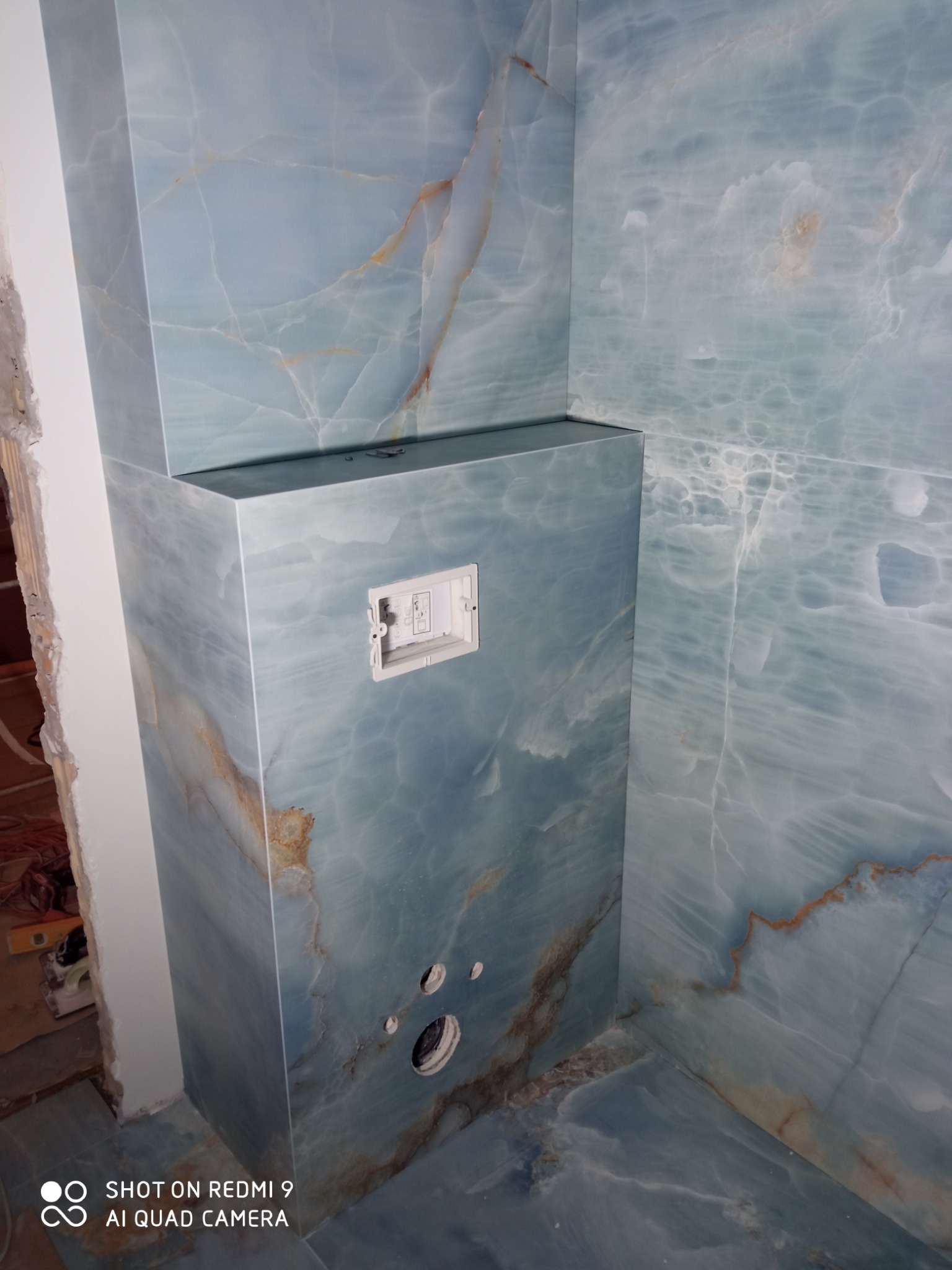 Niebieska mozaika imitująca marmur zdobi obudowę stelaża WC w łazience. Widoczne otwory na rury, biała ramka na przycisk spłukiwania i fragment surowej ściany.