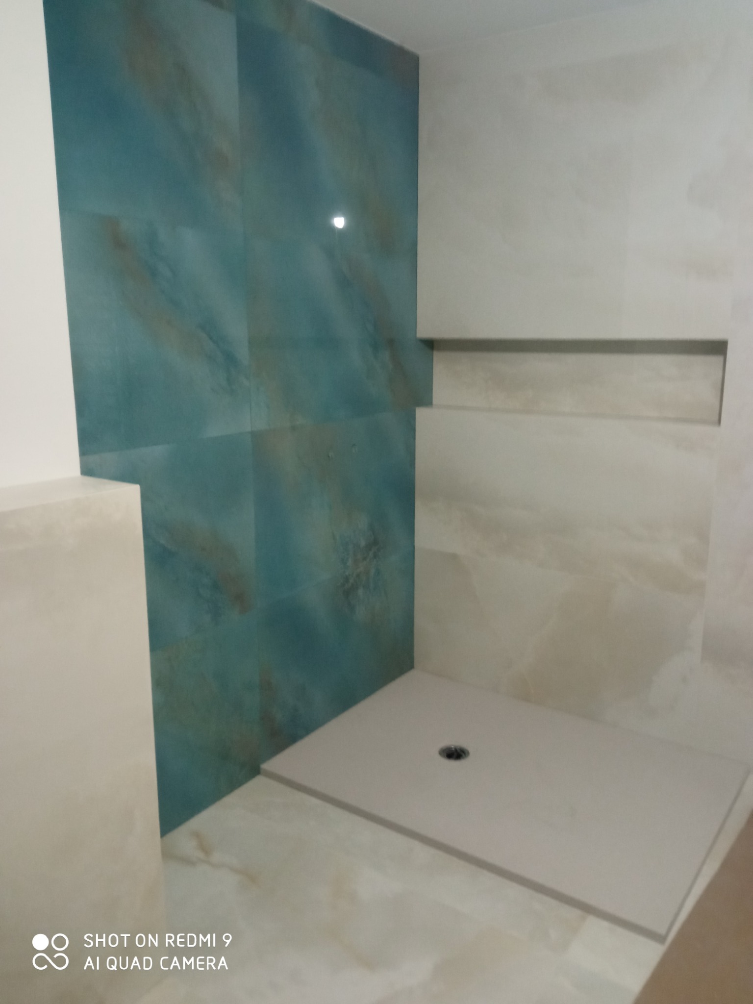 Wykończona łazienka z kabiną typu walk-in, wyłożona płytkami w odcieniach beżu i turkusu. Widoczny odpływ liniowy i wnęka w ścianie. Minimalistyczny design.