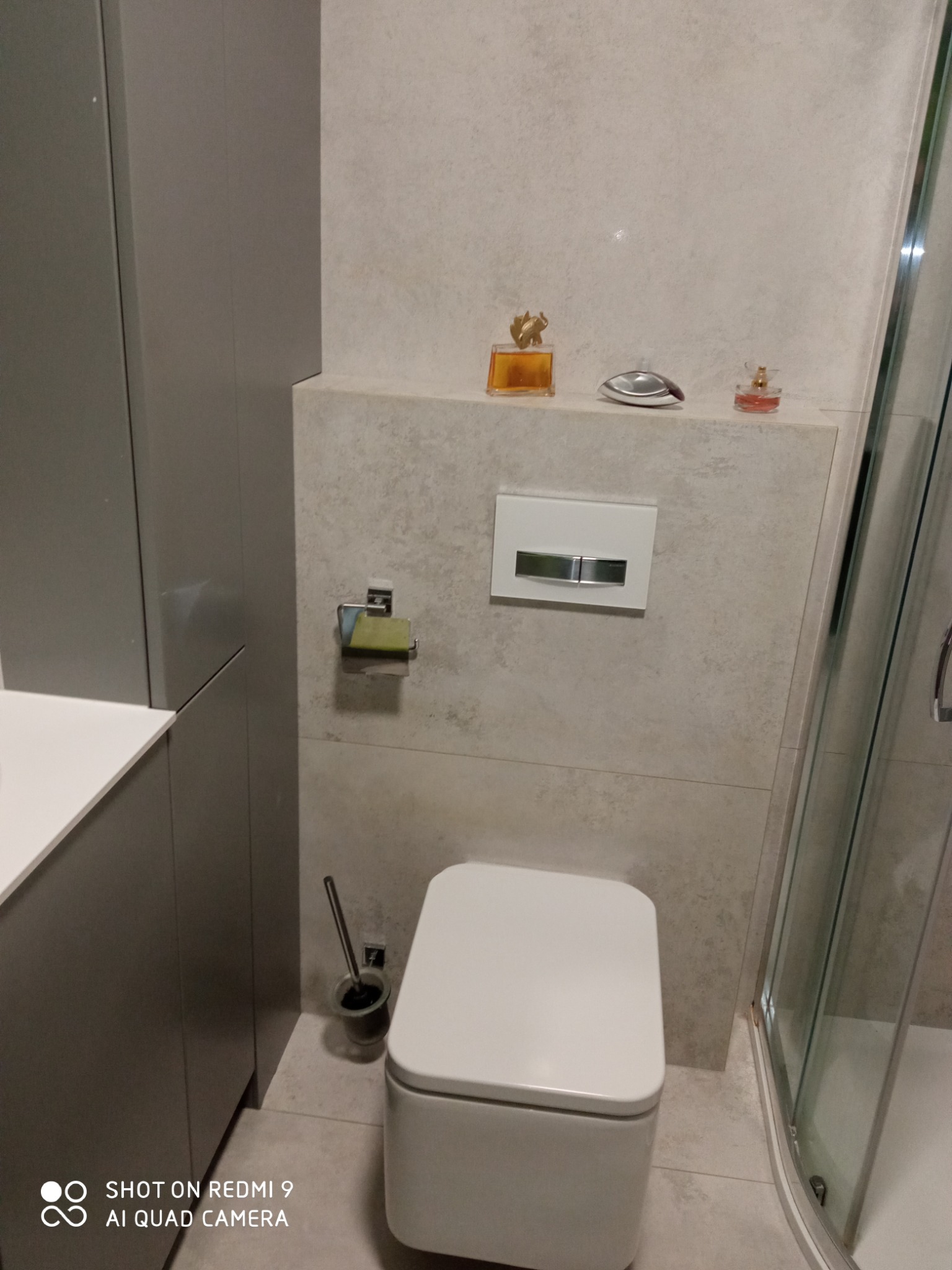 Nowoczesna toaleta w jasnej łazience z szarymi szafkami, widok z góry. Na półce dekoracyjne flakoniki perfum. Minimalistyczny design i neutralna kolorystyka.