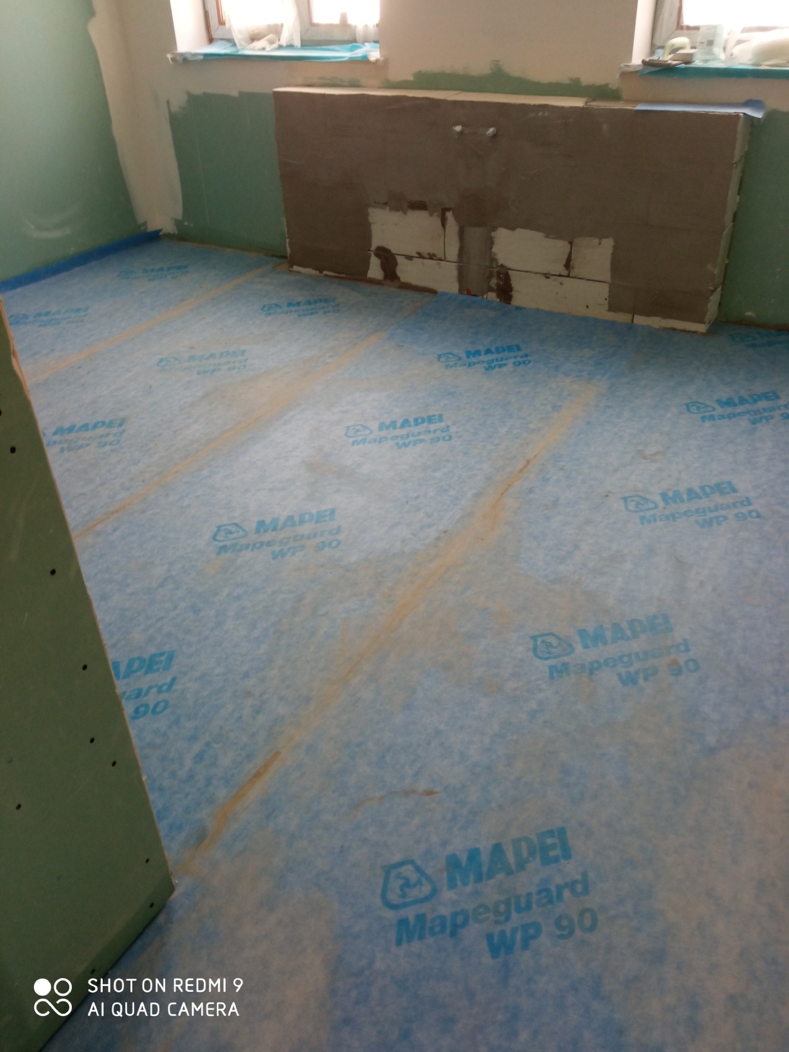 Hydroizolacja podłoża folią Mapeguard WP 90 w łazience przed położeniem płytek. Przygotowanie do remontu, ściany częściowo pokryte klejem. Widoczne okno.