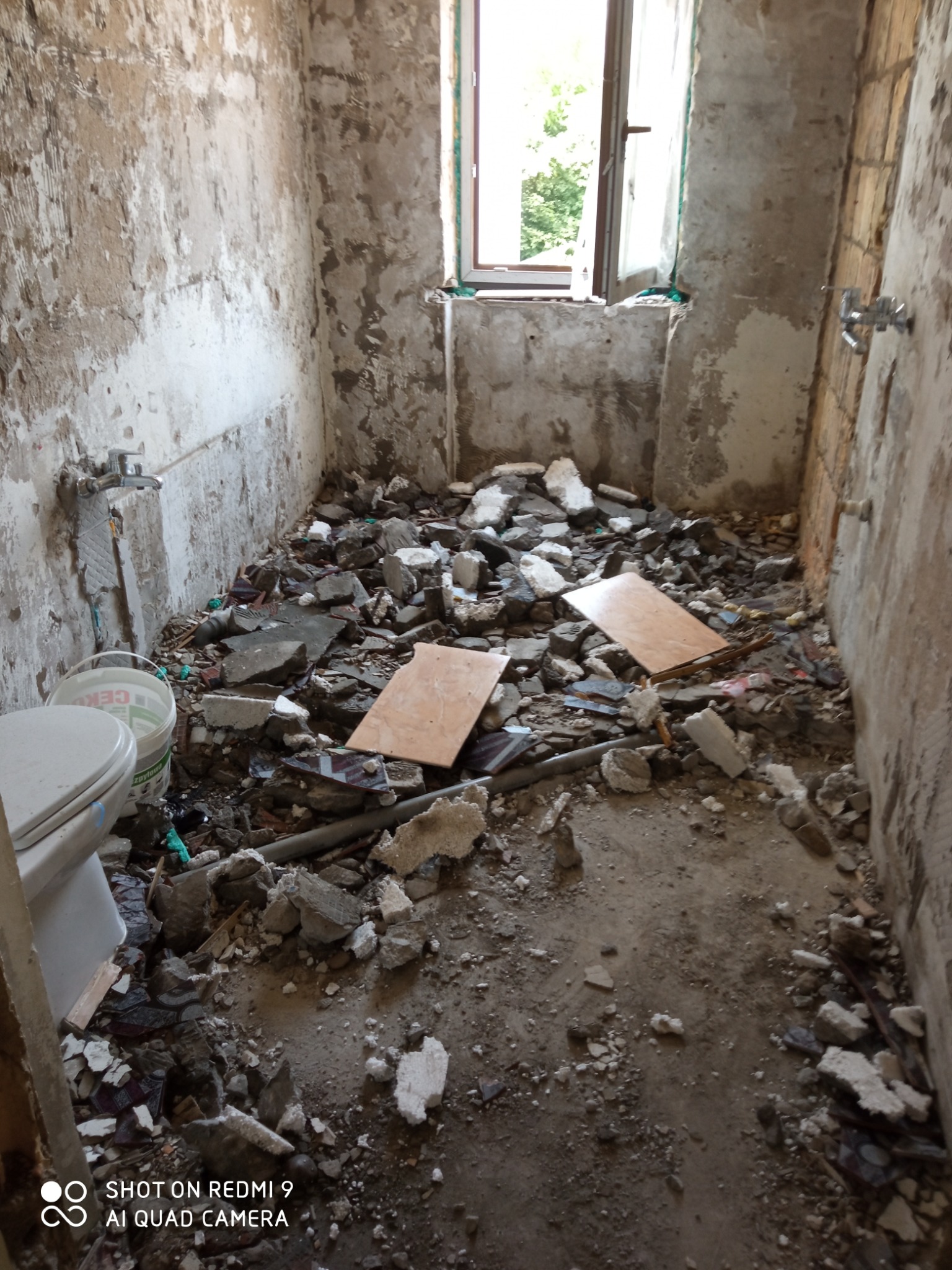 Łazienka w trakcie demontażu: gruz, odłupane płytki, odsłonięte ściany. Widoczne fragmenty toalety i okno z zielenią na zewnątrz. Stan przed remontem.