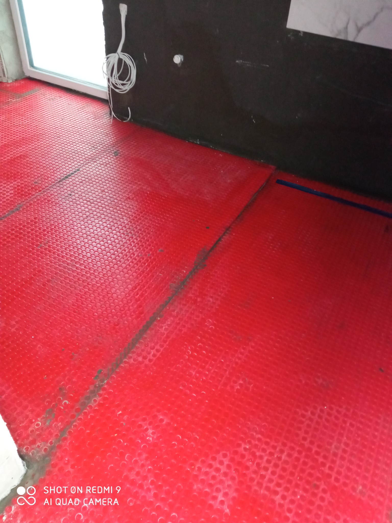 Czerwona mata grzewcza podłogowa z wypustkami, ułożona w rogu pomieszczenia z widocznymi kablami i fragmentem ściany przygotowanej do dalszych prac wykończeniowych.