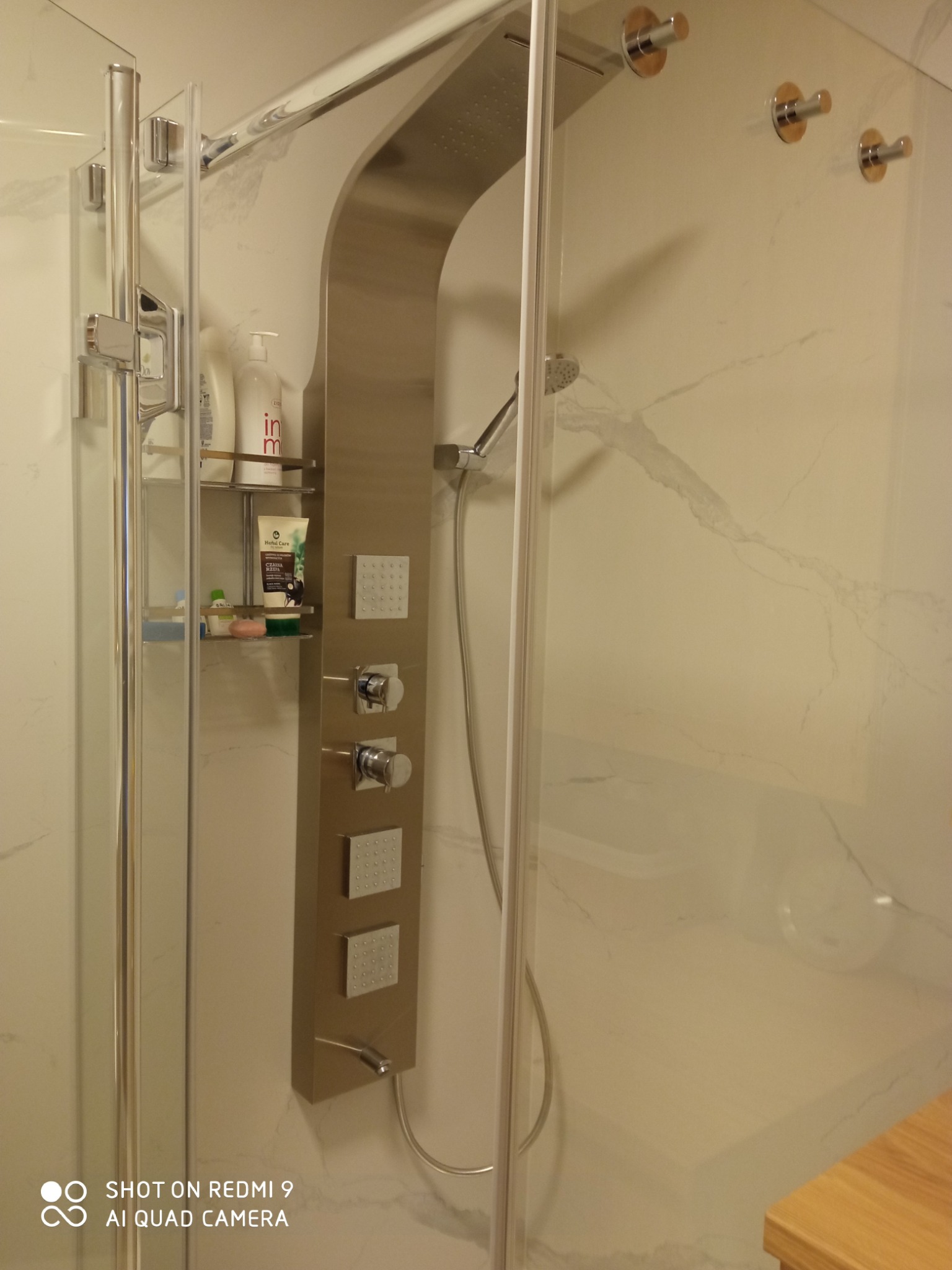 Nowoczesna kabina prysznicowa z panelem natryskowym ze stali nierdzewnej, półką na kosmetyki i marmurowymi ścianami. Widoczne detale wykończenia.