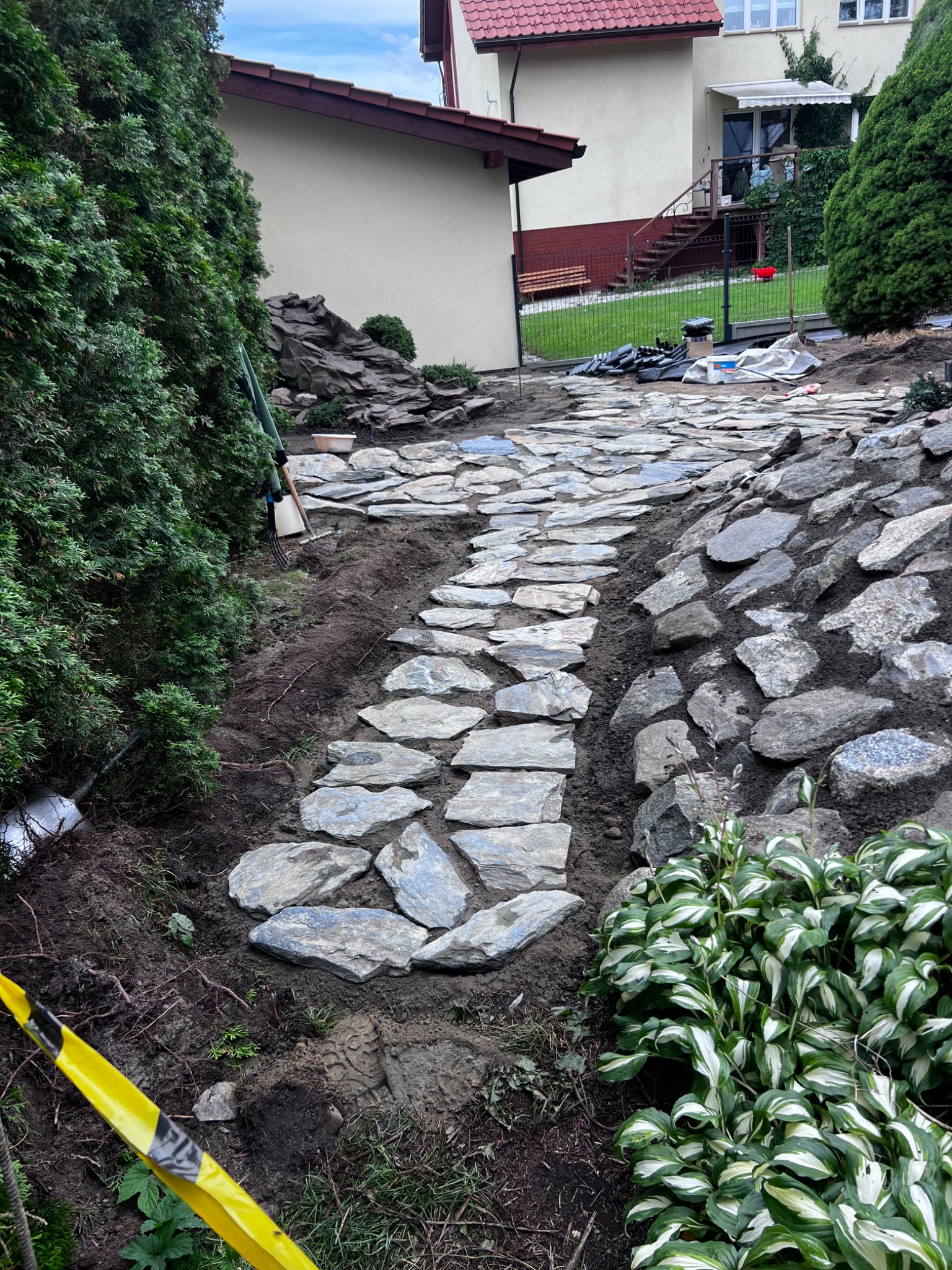 Ścieżka z nieregularnych płyt kamiennych w ogrodzie, otoczona zielenią i kamieniami, prowadząca w górę zbocza w kierunku domu.