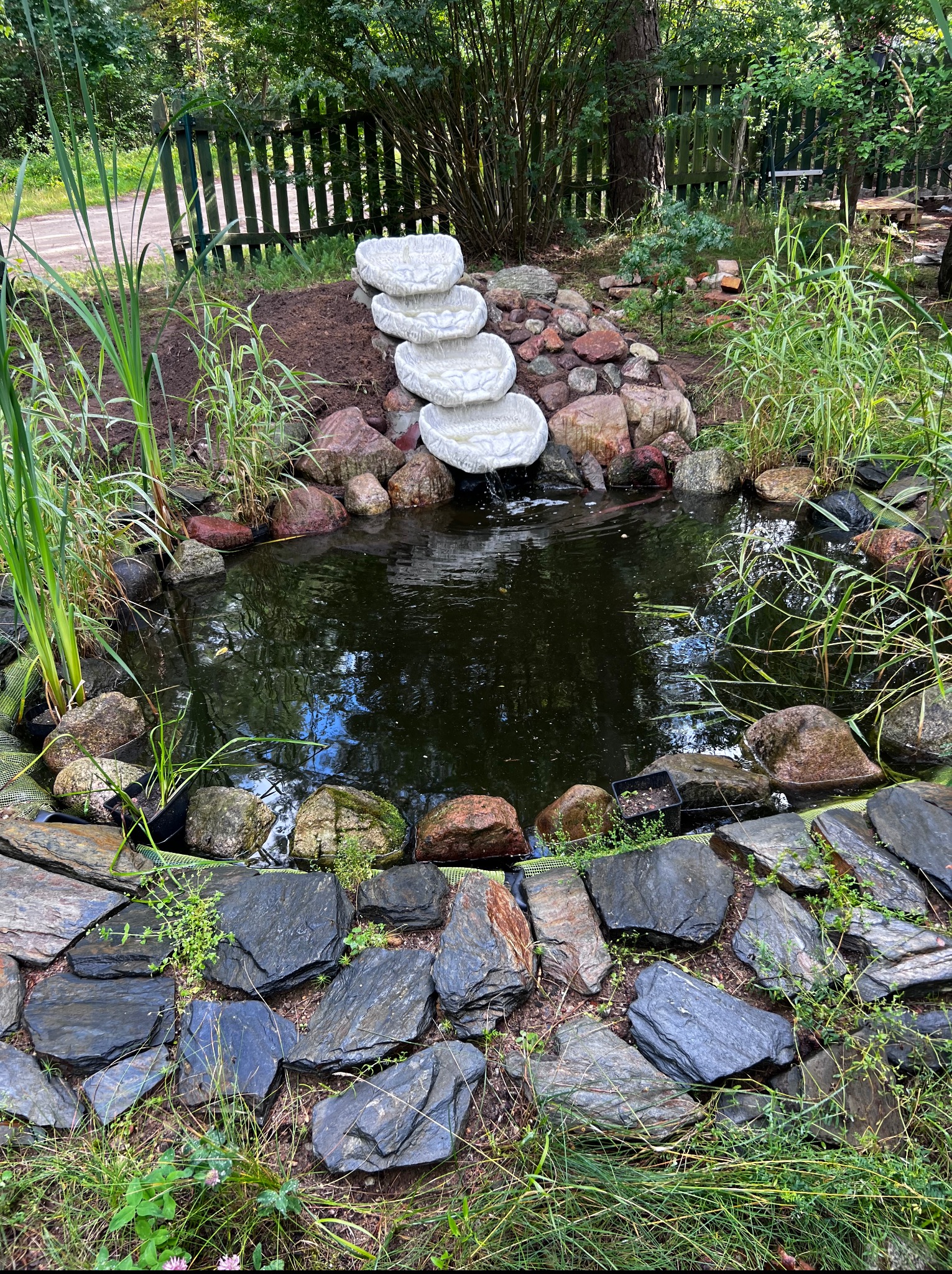 Oczko wodne z kamienną kaskadą i obrzeżem z płaskich kamieni, otoczone zielenią. Woda odbija niebo, tworząc spokojny krajobraz ogrodu.