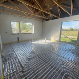 Instalacja ogrzewania podłogowego w nowym domu w Markach. Białe rury ułożone na folii, widok na surowy strop i okna z widokiem na zewnątrz.