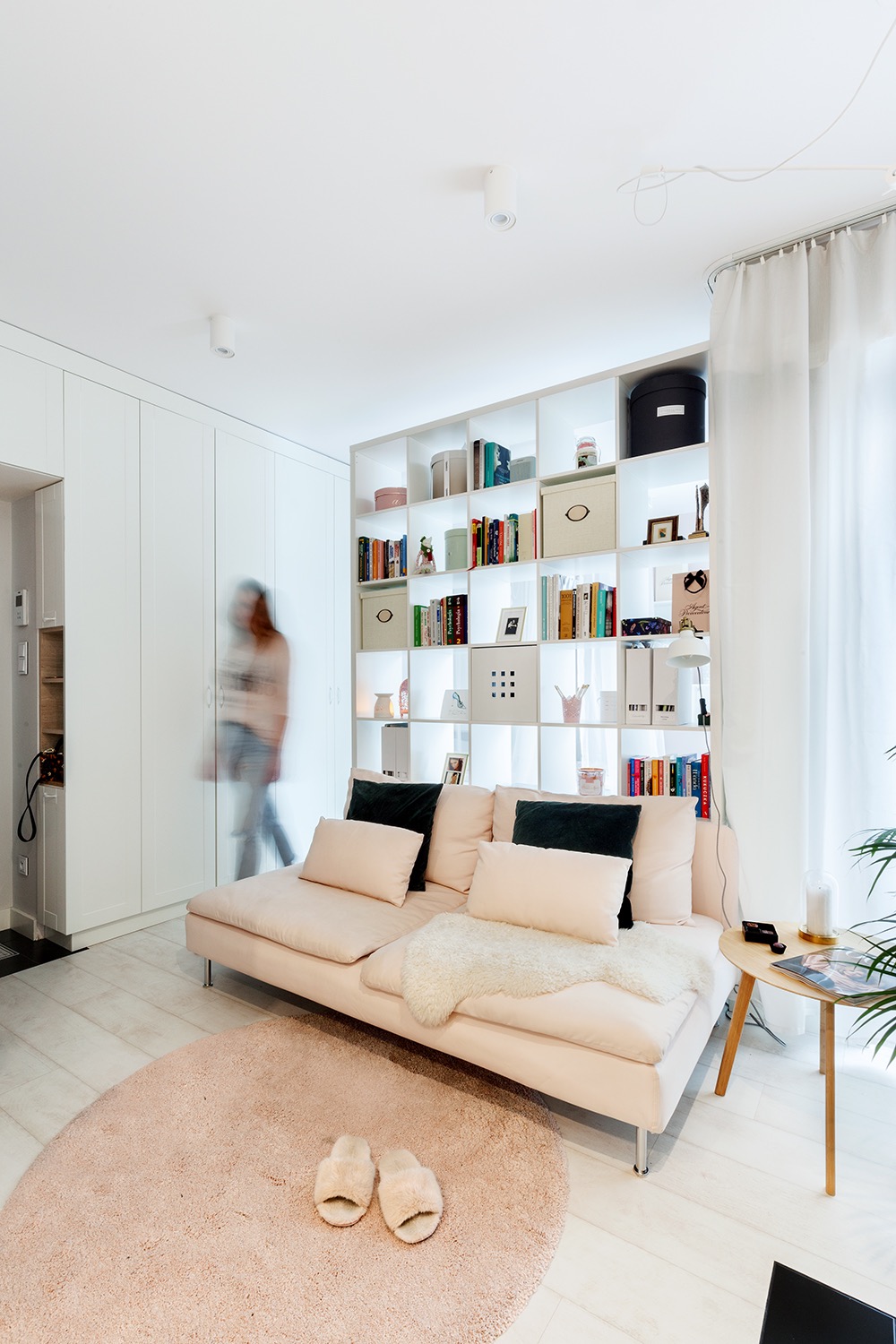 Jasne wnętrze z pastelową sofą, regałem z podświetleniem i rozmytą sylwetką osoby, tworzące przytulną, minimalistyczną przestrzeń.