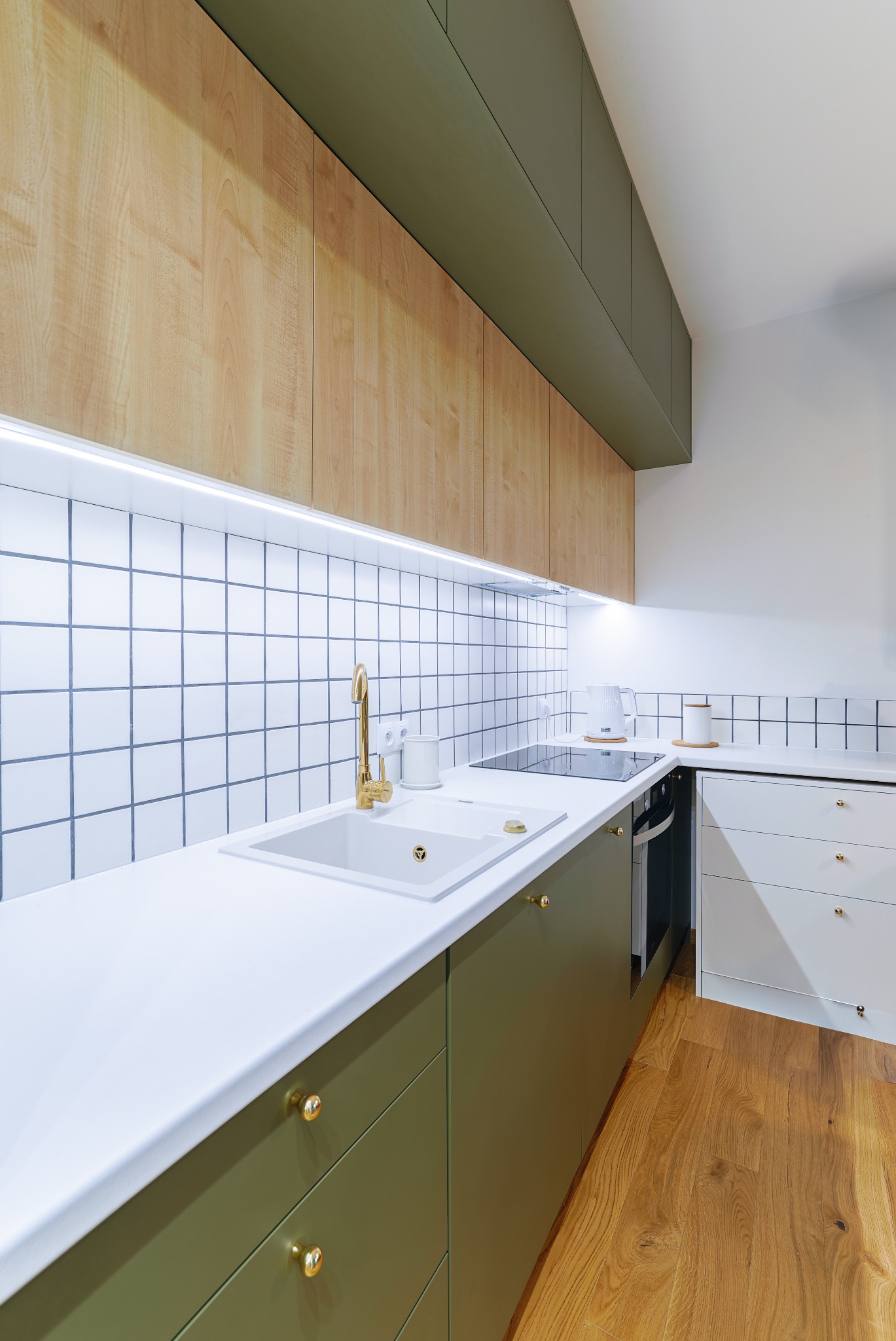 Nowoczesna kuchnia z drewnianymi szafkami i oliwkowymi frontami, biały blat, złota armatura i płytki w kratkę. Podświetlenie LED podszafkowe.
