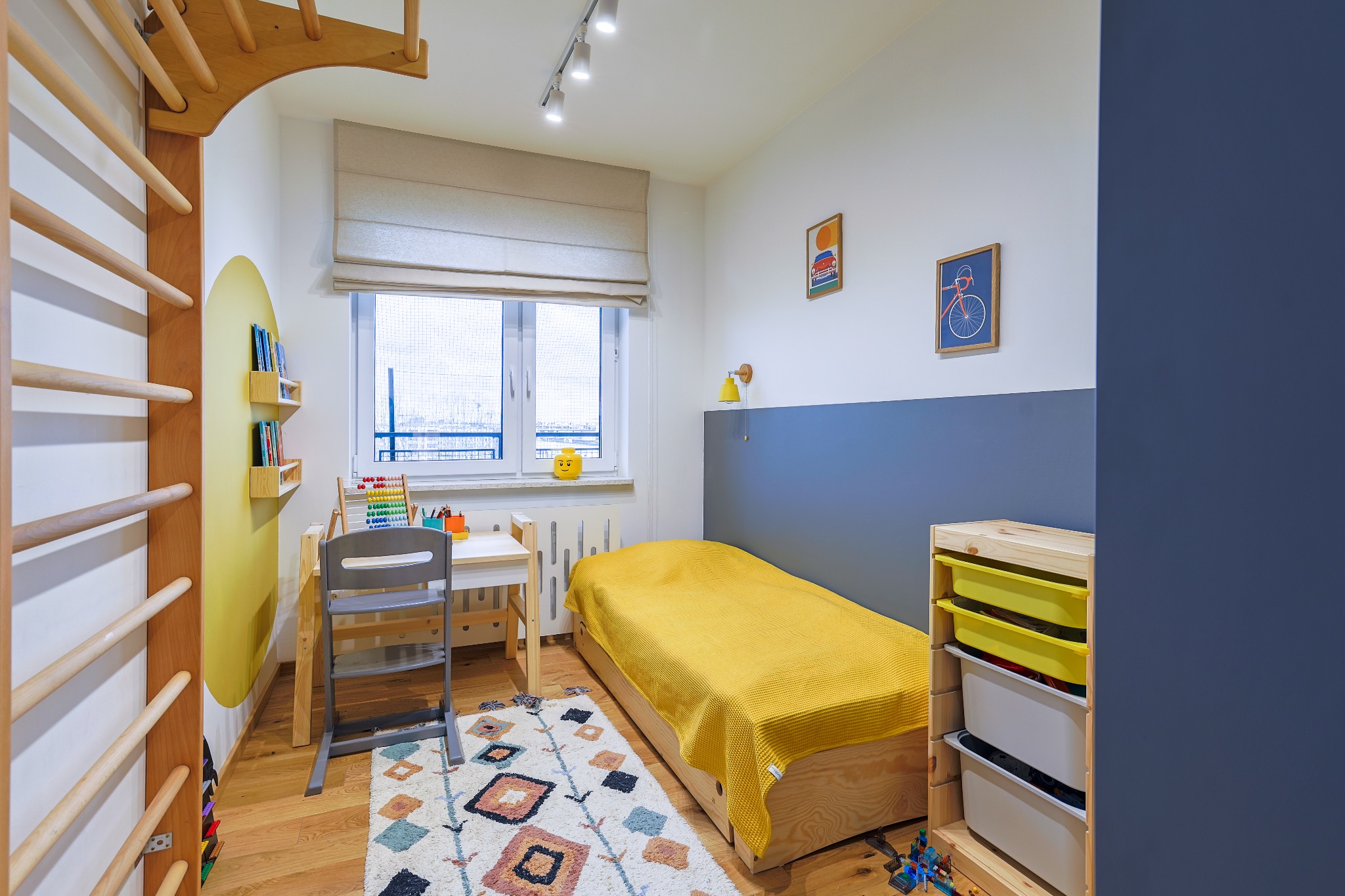 Pokój dziecięcy w Markach: drewniane drabinki gimnastyczne, biurko z krzesłem, łóżko z żółtą narzutą, kolorowe dekoracje i dywan w geometryczne wzory.