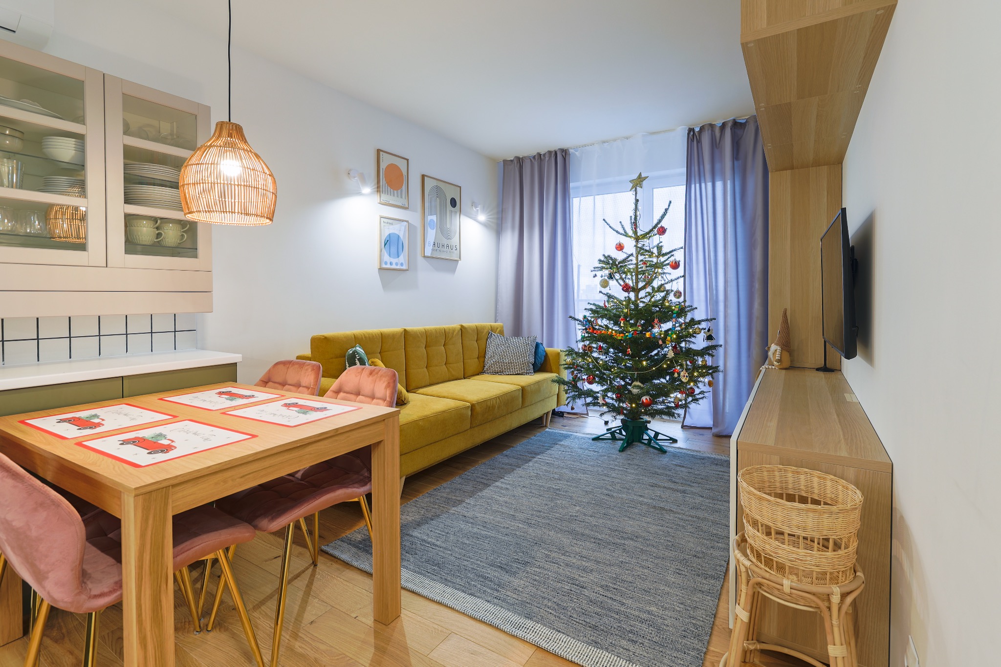 Jasne wnętrze z żółtą sofą, choinką i drewnianym stołem z różowymi krzesłami. W tle minimalistyczne grafiki i zasłony w odcieniach fioletu.