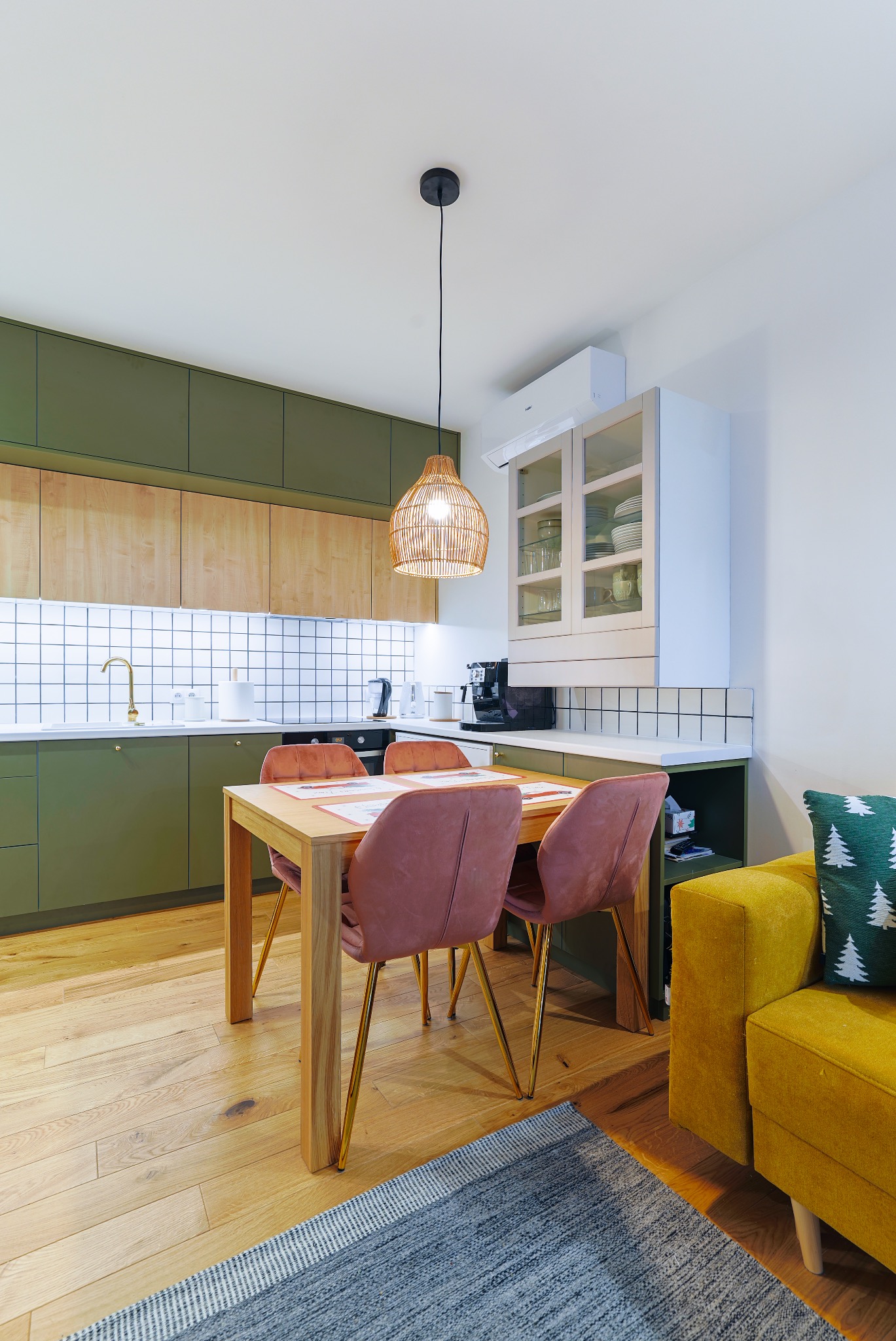 Nowoczesna kuchnia z oliwkowymi szafkami, drewnianym blatem i złotą armaturą. Stół z różowymi krzesłami i lampa z wikliny dodają wnętrzu przytulności.