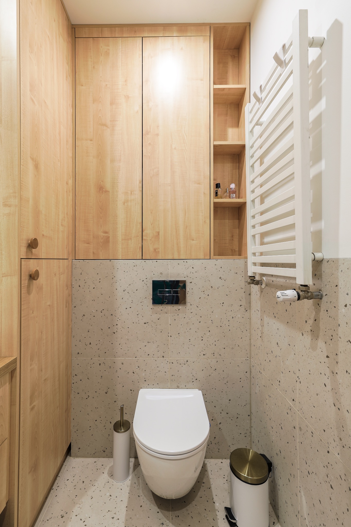 Nowoczesna łazienka z drewnianą zabudową, mozaikowymi płytkami i białym grzejnikiem drabinkowym. Minimalistyczny design z akcentami w odcieniach drewna i złota.