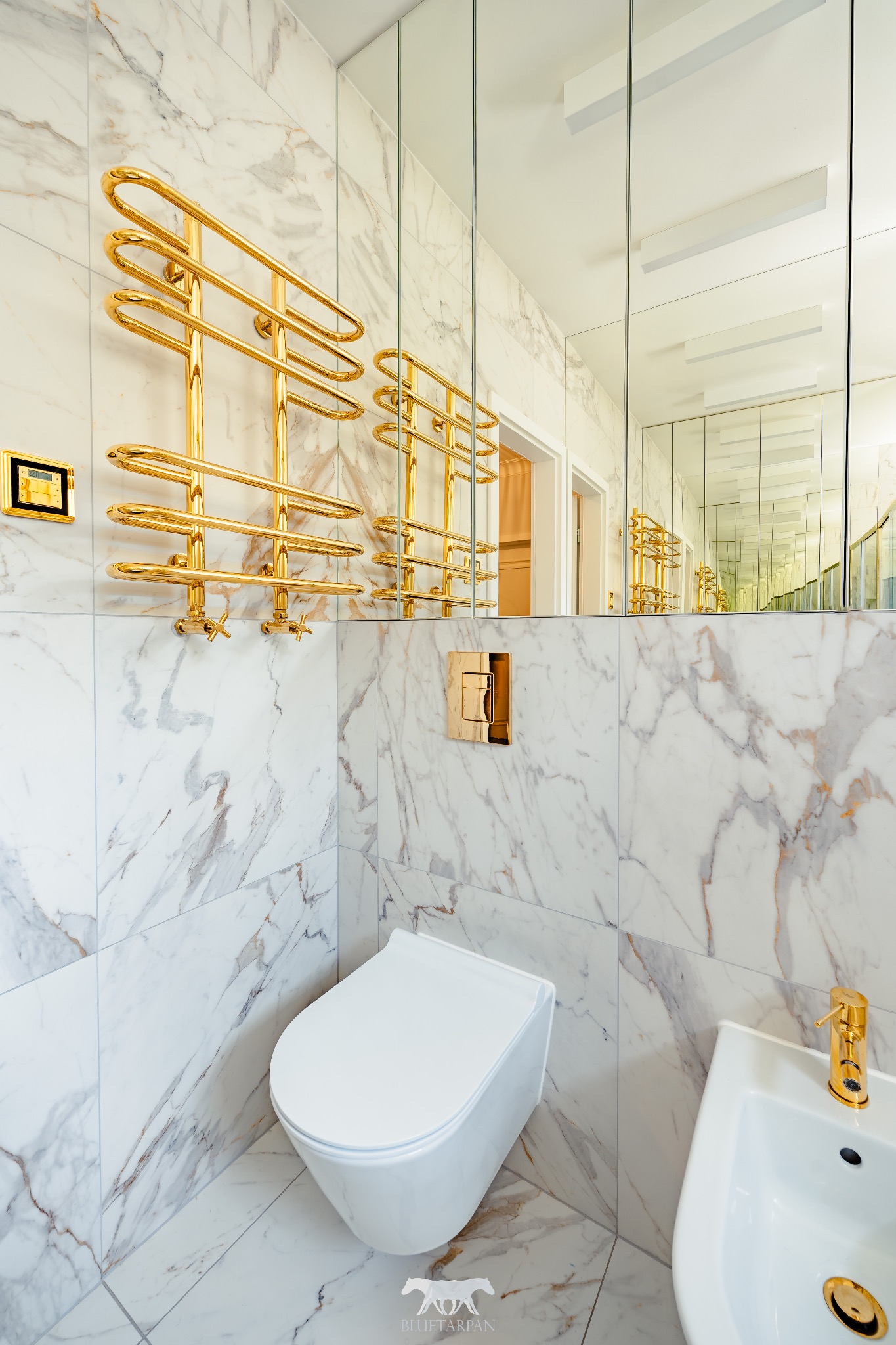 Luksusowa łazienka z marmurowymi płytkami, złotym grzejnikiem drabinkowym i armaturą. Lustro optycznie powiększa przestrzeń. Biała toaleta i bidet.