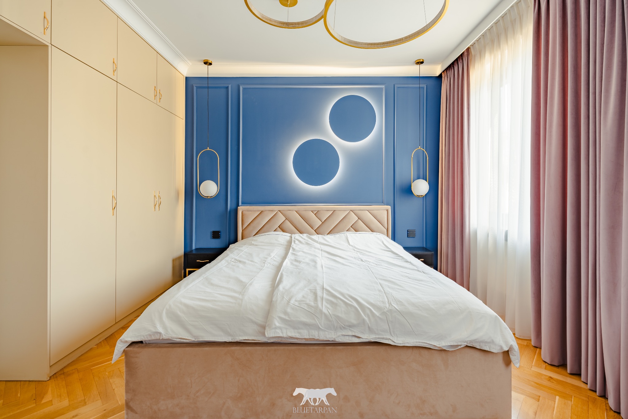 Nowoczesna sypialnia: błękitna ściana z oświetleniem, beżowe łóżko i szafa, parkiet. Złote lampy i dekoracje dodają elegancji.