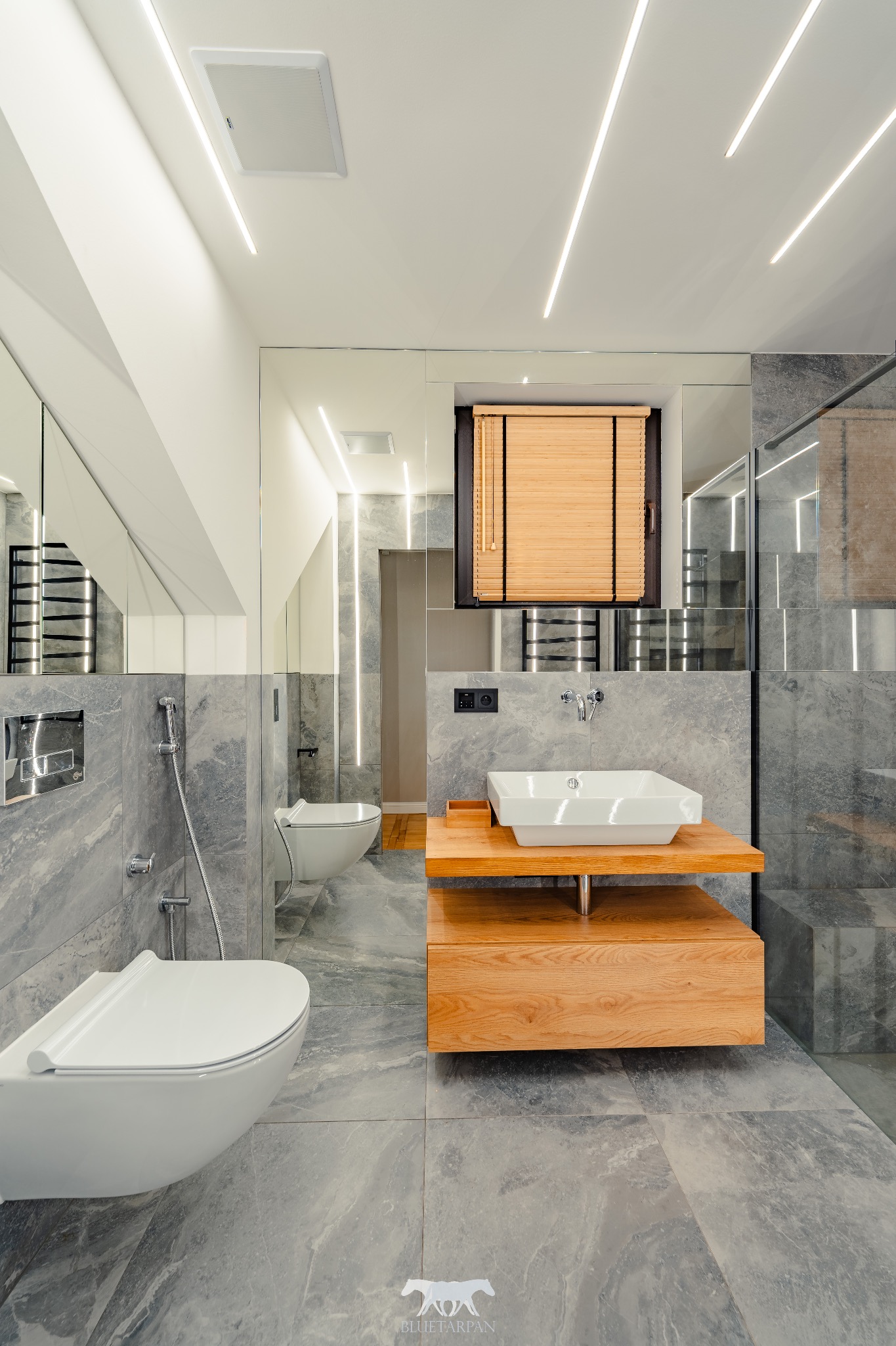 Stylowa łazienka z szarymi płytkami imitującymi marmur, podwieszaną toaletą, drewnianą szafką pod umywalką i oświetleniem LED w suficie.