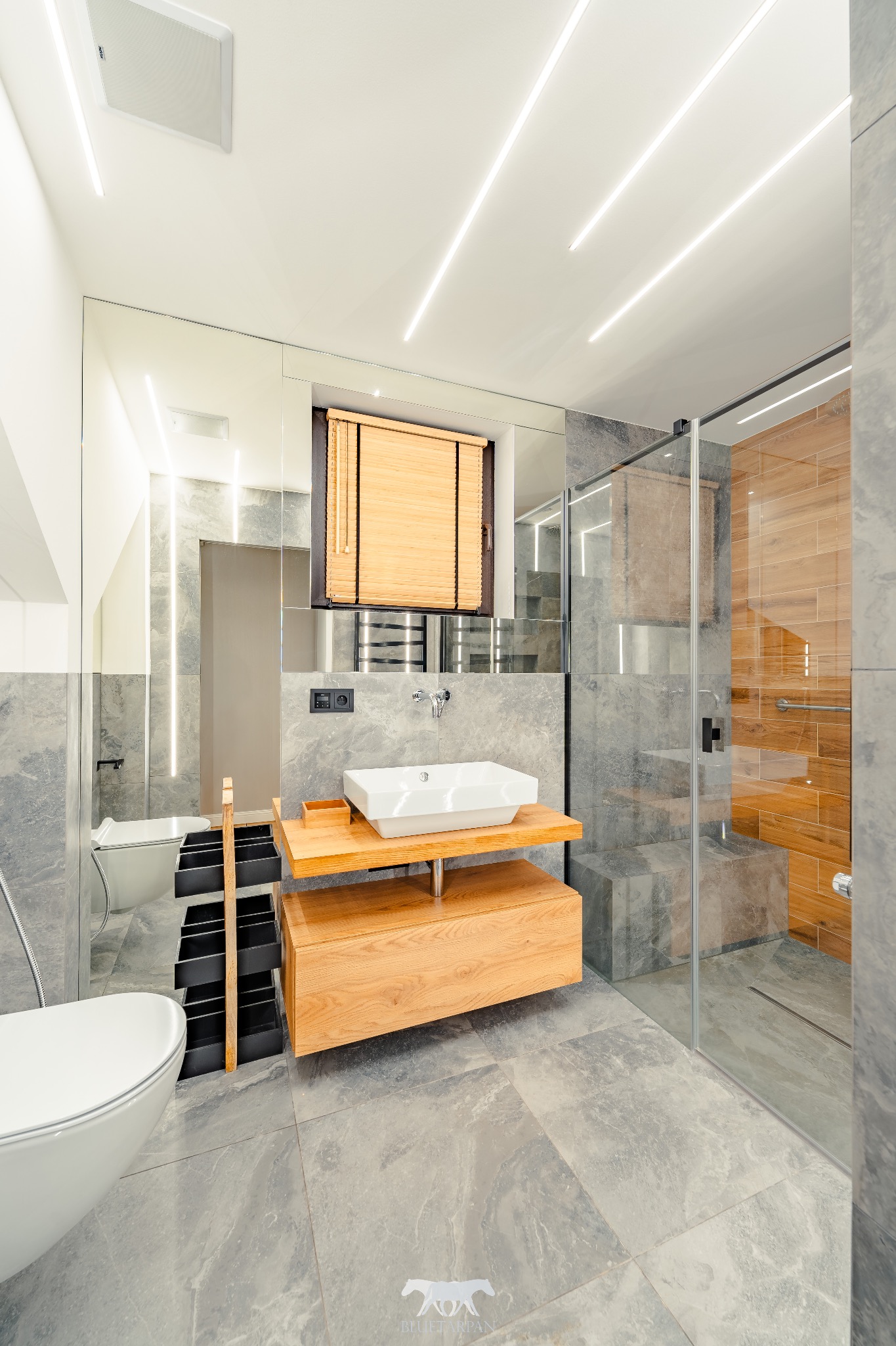 Nowoczesna łazienka z szarymi płytkami, drewnianą szafką pod umywalką i kabiną prysznicową z drewnianą ścianą. Oświetlenie ledowe podkreśla minimalistyczny design.