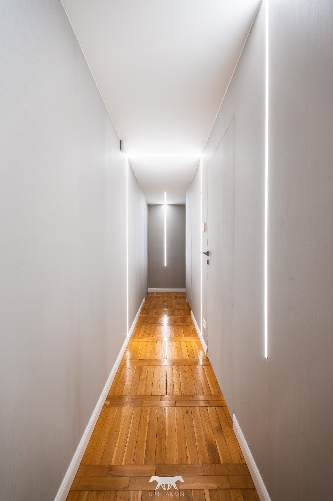 Wąski korytarz z parkietem i pionowymi pasami światła LED na ścianach, tworzącymi nowoczesny efekt oświetleniowy. Minimalistyczny design i ciepła barwa drewna.
