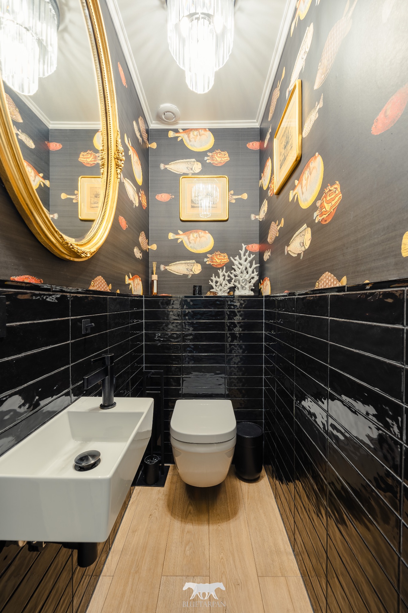 Nowoczesna toaleta z czarnymi, błyszczącymi płytkami ułożonymi poziomo, drewnianą podłogą i tapetą w ryby. Złote lustro i dekoracje dodają elegancji.