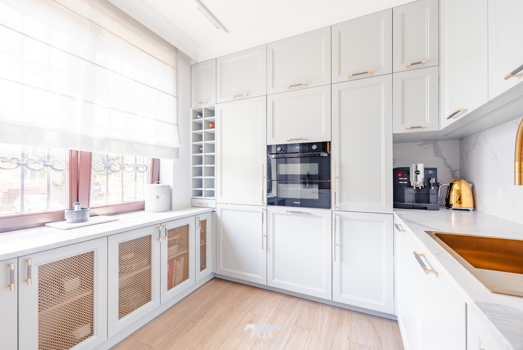 Jasna, elegancka kuchnia z białymi szafkami, złotymi akcentami, drewnianą podłogą i marmurowym blatem. Widoczne okno z roletami i ekspres do kawy.