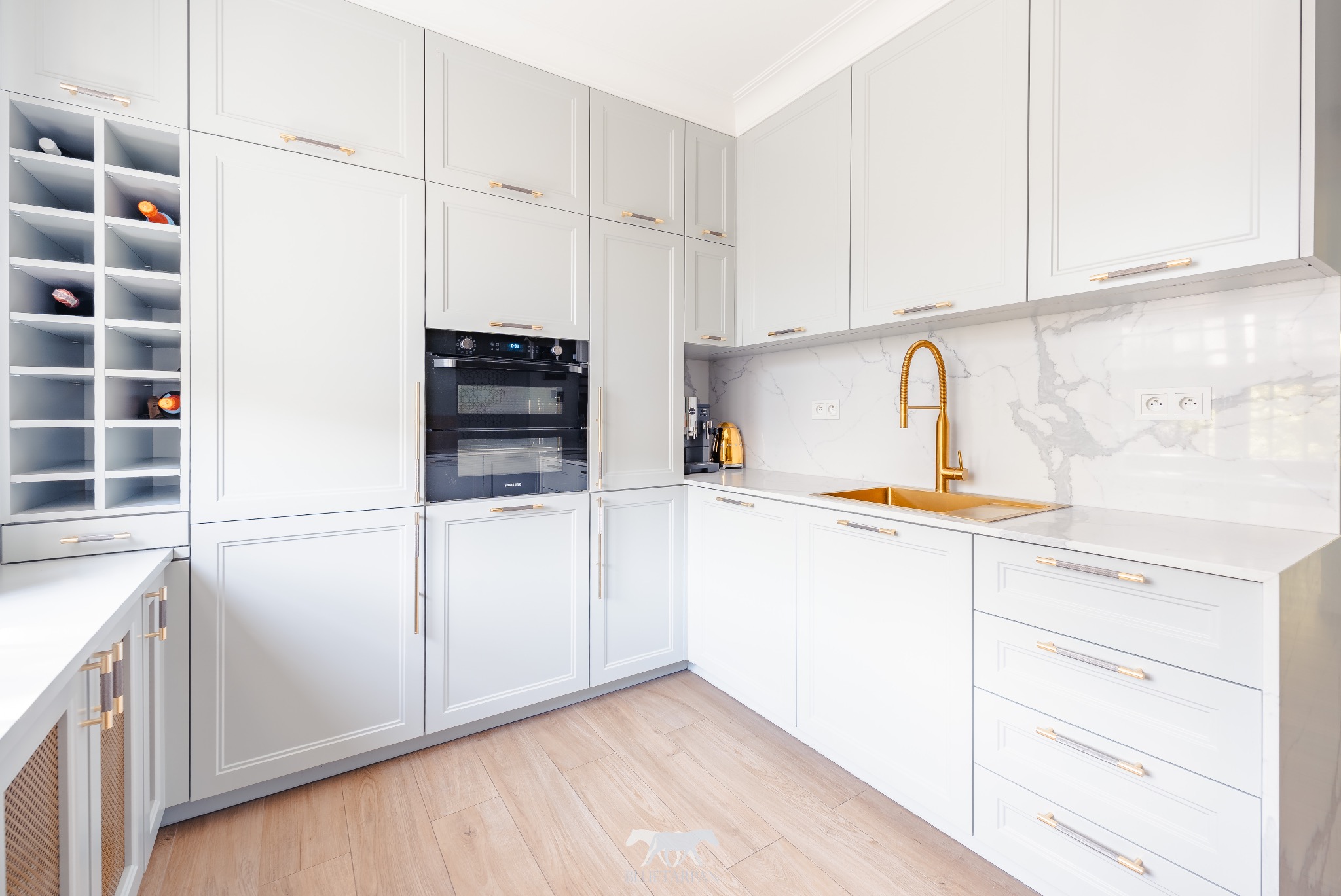 Elegancka, jasnoszara kuchnia z złotymi akcentami: bateria, zlew i uchwyty. W tle marmurowy backsplash i wbudowany regał na wino. Parkiet dodaje ciepła.