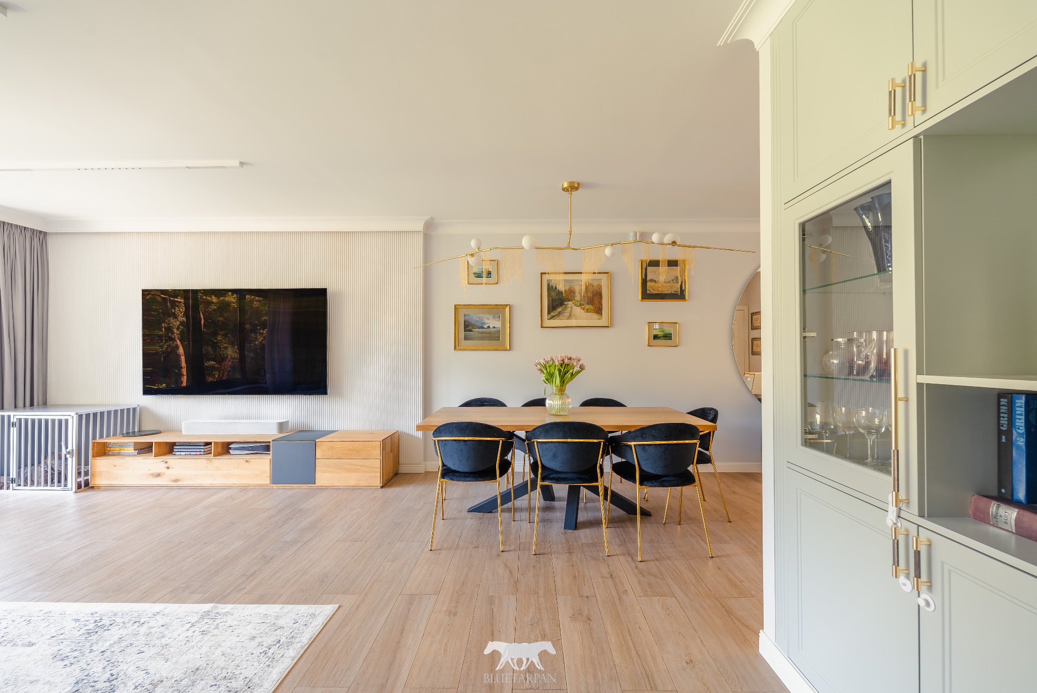 Nowoczesne wnętrze: drewniana podłoga, stół z czarnymi krzesłami, telewizor na ścianie z lamelami. Uwagę przyciąga złoty żyrandol i przeszklona witryna.