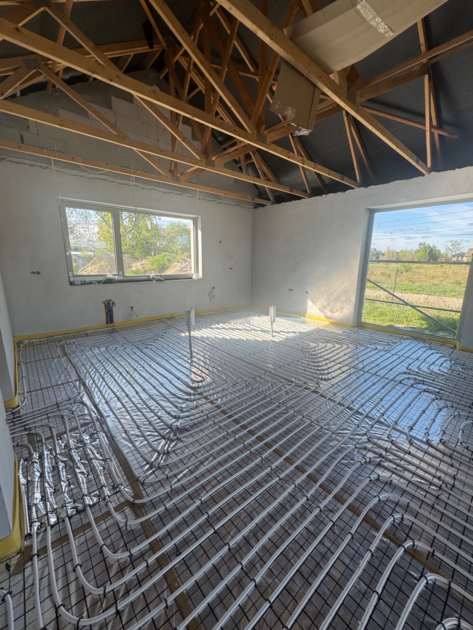 Instalacja ogrzewania podłogowego w nowym domu w Markach. Białe rury ułożone na folii, widok na surowy strop i okna z widokiem na zewnątrz.