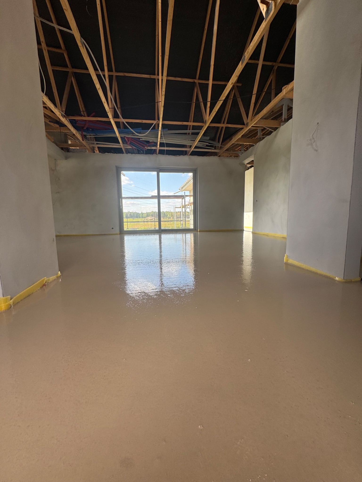 Wylany, mokry podkład podłogowy w surowym wnętrzu domu z odsłoniętą konstrukcją dachu i oknem z widokiem na zewnątrz. Taśma zabezpieczająca przy ścianach.