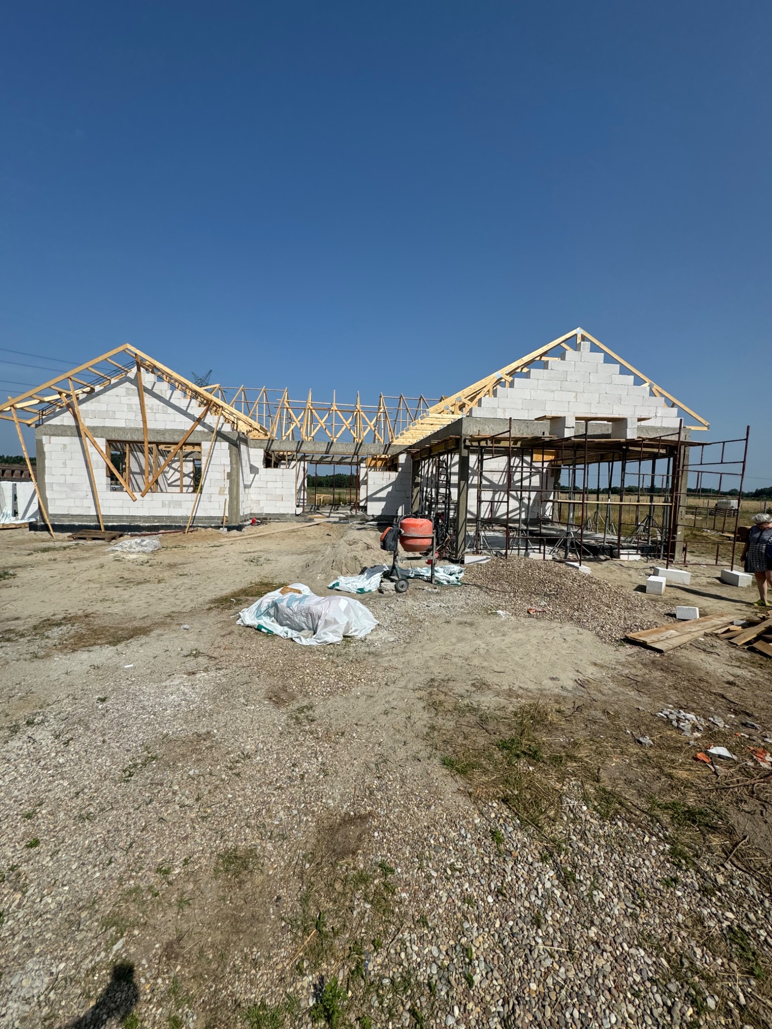 Budowa domu z białych bloczków, widoczne drewniane elementy konstrukcji dachu, rusztowania, betoniarka i materiały budowlane na placu budowy w słoneczny dzień.