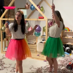 Dwie animatorki w kolorowych tiulowych spódnicach prezentują sztuczki z talerzem i pałeczką na tle dekoracji urodzinowej z balonami i drewnianym domkiem.