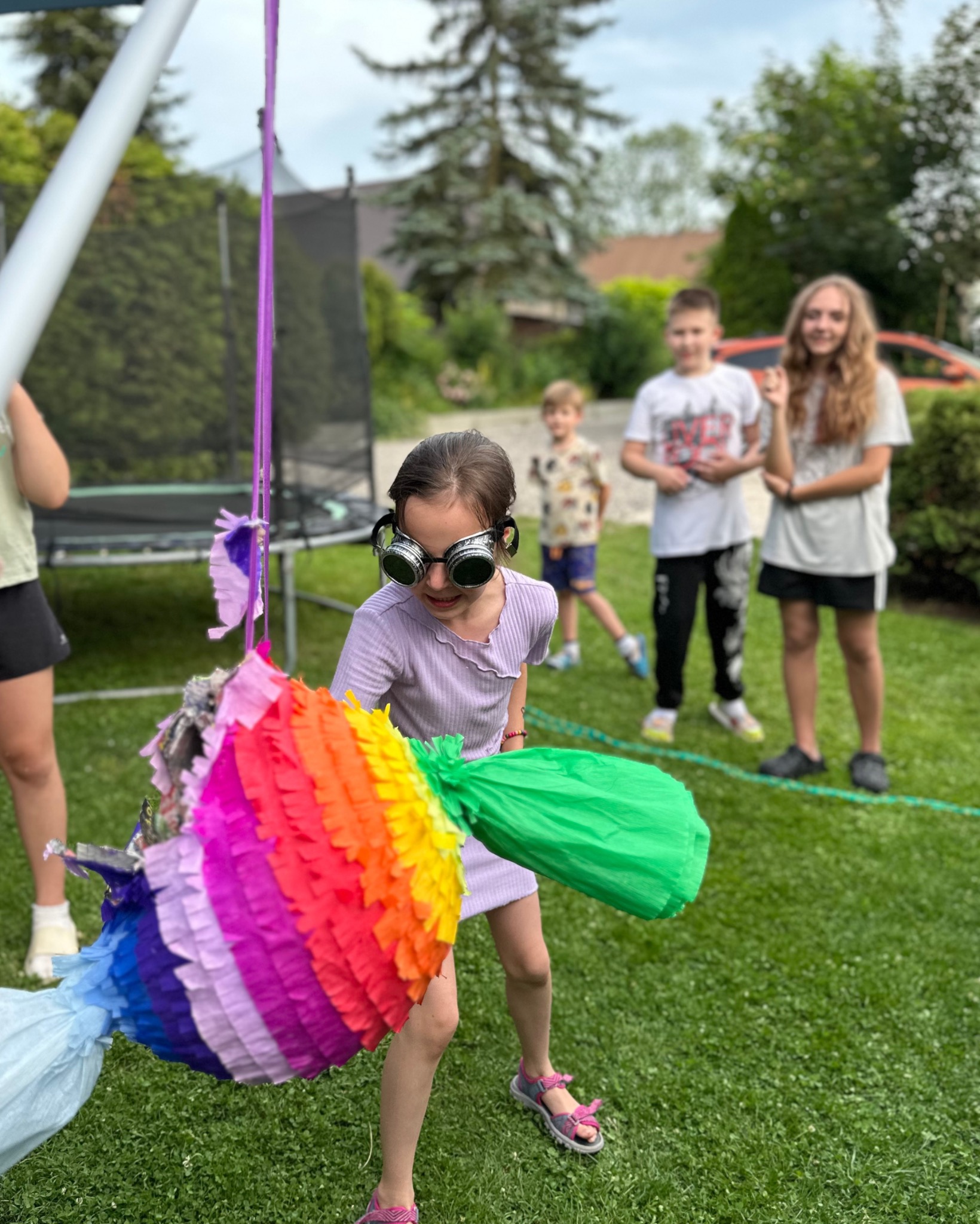 Dziewczynka w śmiesznych okularach uderza w kolorową piniatę na tle zielonego trawnika i obserwujących dzieci. Zabawa na imprezie plenerowej.