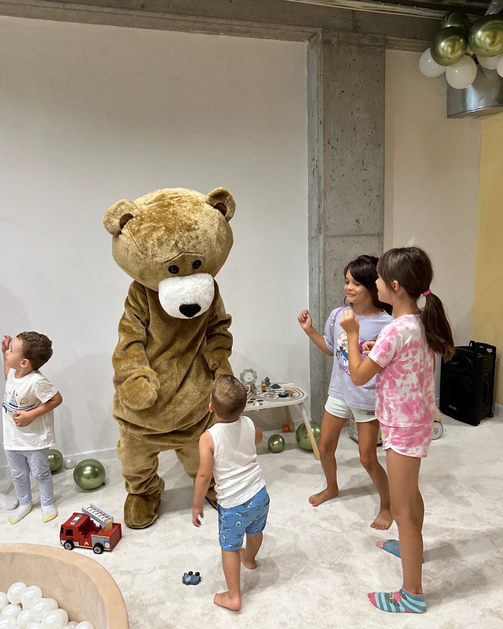 Animator w kostiumie misia bawi dzieci w sali zabaw z basenem z kulkami i dekoracjami z balonów. W tle widoczny betonowy filar i mały stolik z zabawkami.