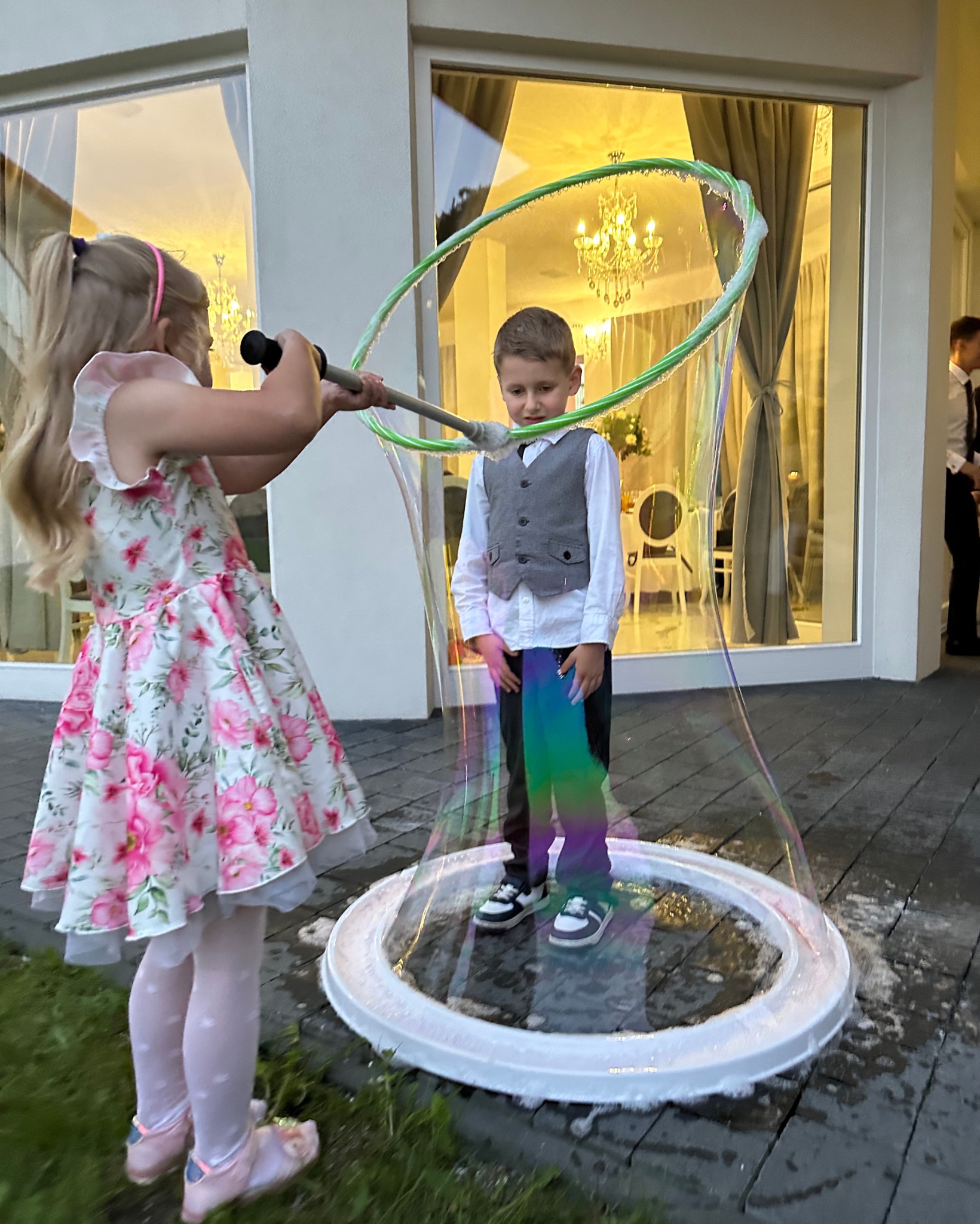 Dziewczynka robi ogromne bańki mydlane, w których stoi chłopiec w eleganckiej kamizelce, na tle jasnego budynku z oknami.