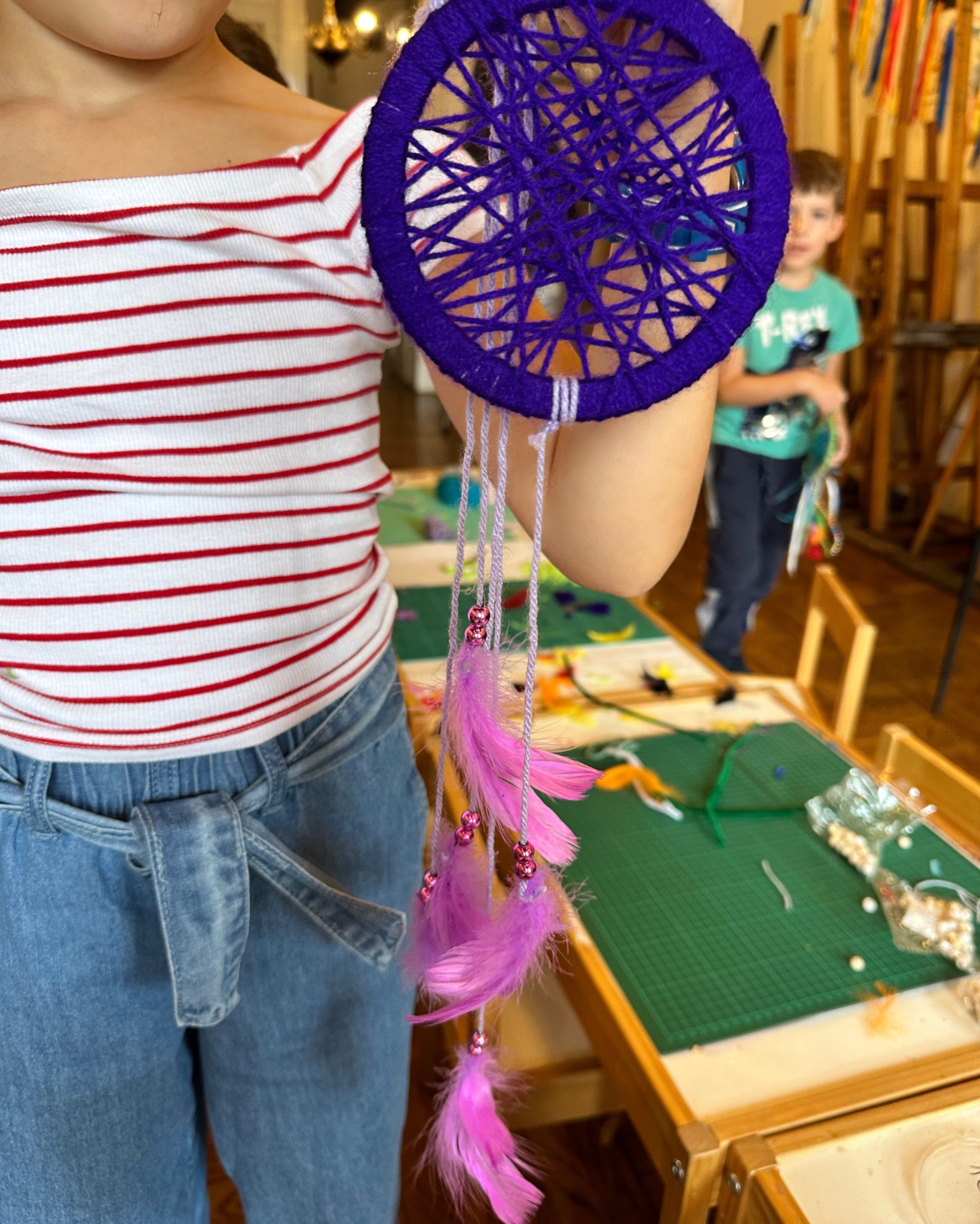 Dziecko prezentuje ręcznie robiony fioletowy łapacz snów z piórami, wykonany podczas warsztatów, na tle stołu z materiałami do rękodzieła i zarysów postaci.