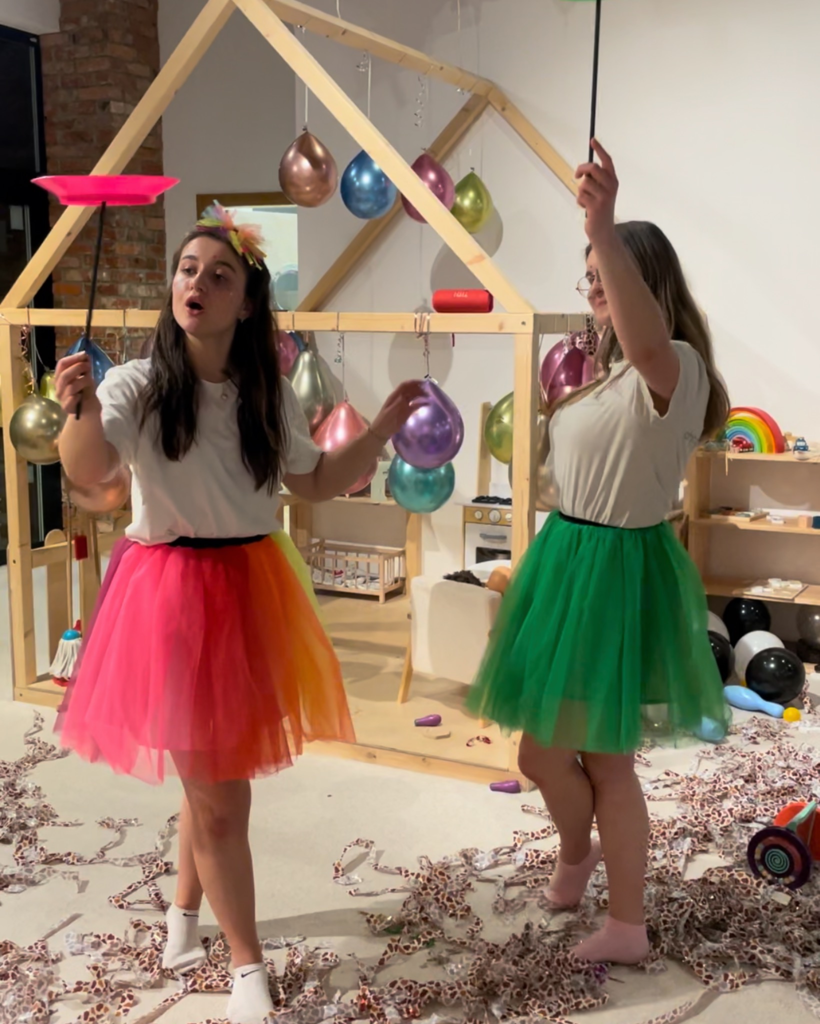 Dwie animatorki w kolorowych tiulowych spódnicach prezentują sztuczki z talerzem i pałeczką na tle dekoracji urodzinowej z balonami i drewnianym domkiem.