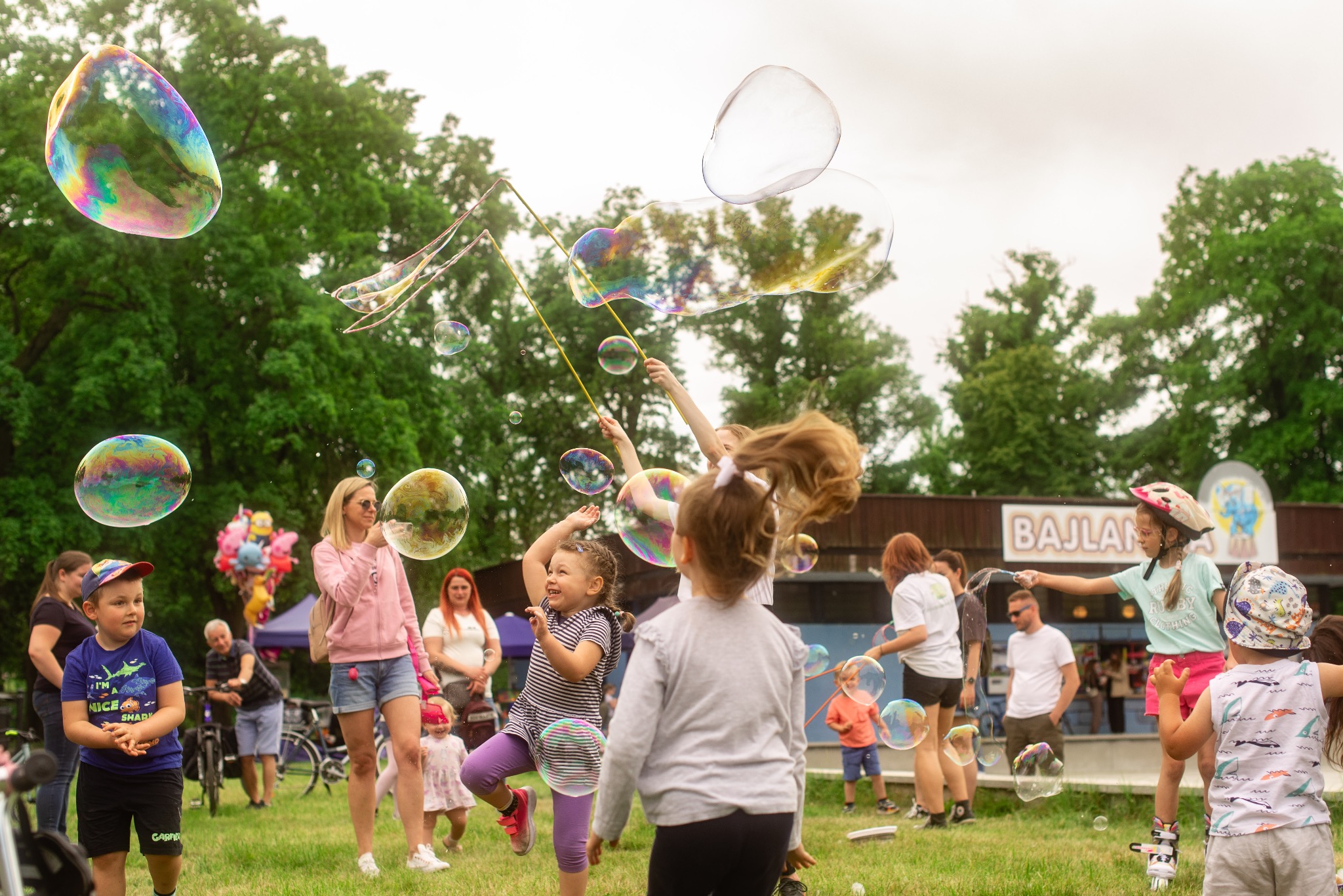Radosne dzieci bawią się na trawie, puszczając ogromne bańki mydlane, z budynkiem w tle. Scena pełna śmiechu i ruchu, idealna na imprezę plenerową.