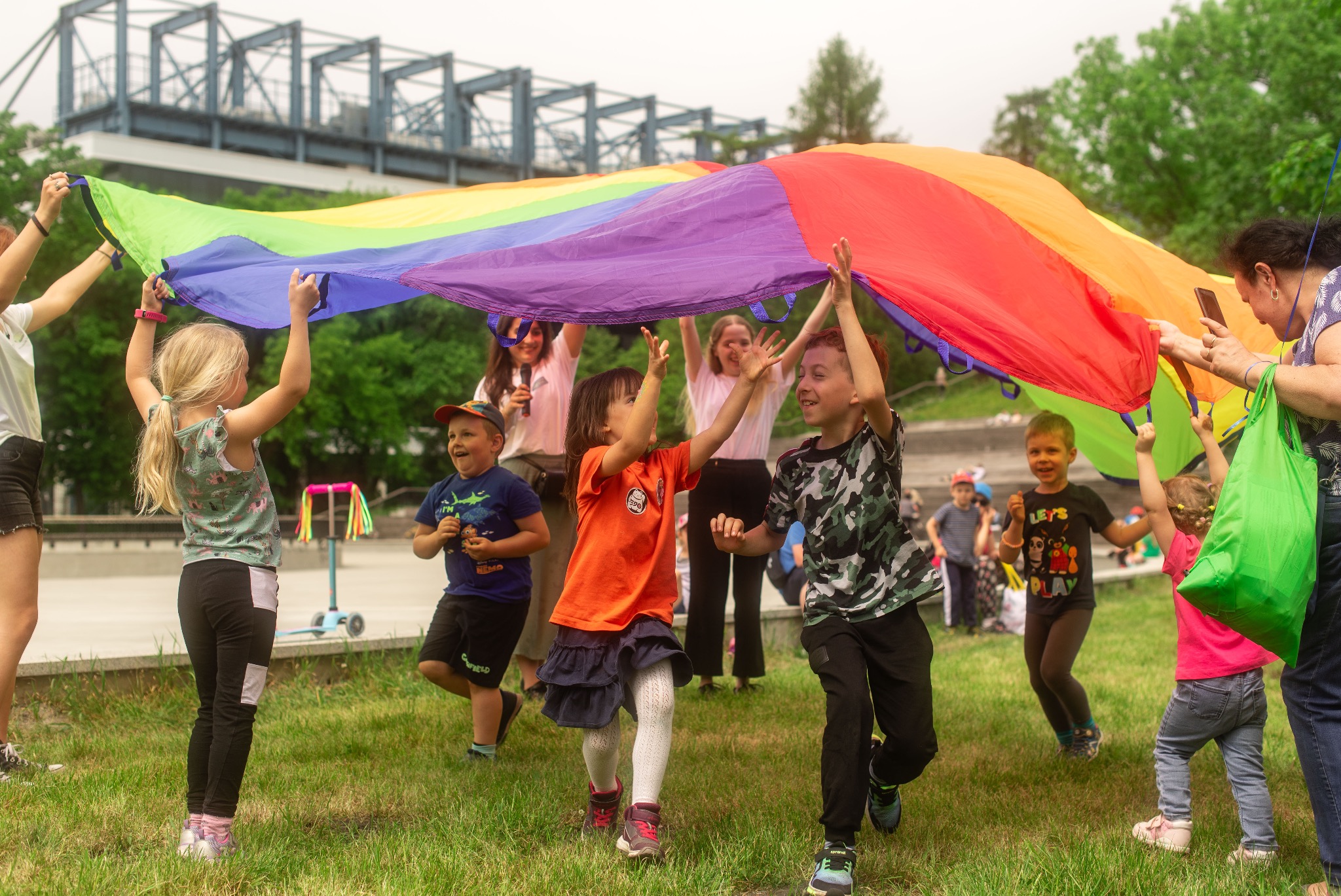 Radosne dzieci bawią się pod tęczową płachtą na zielonej trawie, w tle zarys konstrukcji mostu. Uczestnicy animacji podczas eventu.