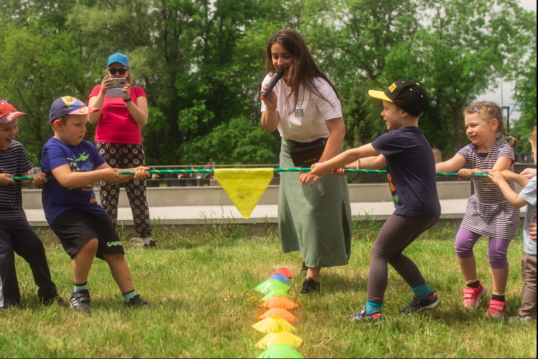 Dzieci biorące udział w przeciąganiu liny na trawie, prowadzone przez animatorkę z mikrofonem. W tle osoba robiąca zdjęcia telefonem. Kolorowe pachołki wyznaczają trasę zabawy.