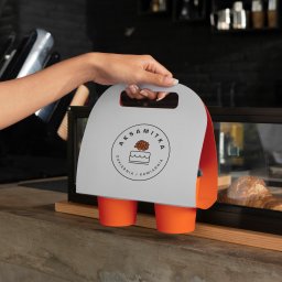 Julita Saliwon Graphic Designer - Dłoń trzyma papierowy uchwyt z logo 'Aksamitka' (cukiernia i kawiarnia) i dwoma pomarańczowymi kubkami na tle lady z croissantem w witrynie.