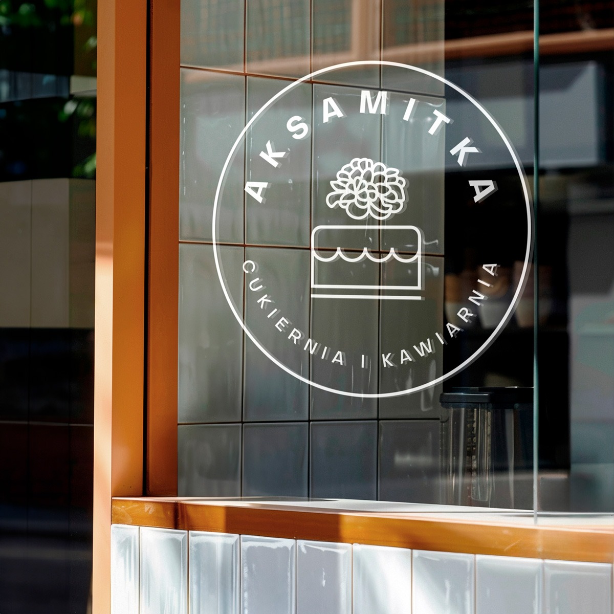 Grafika na szkle: logo kawiarni „Aksamitka” z motywem ciasta i kwiatu, otoczone napisem. Widoczne płytki ceramiczne w tle i elementy wystroju wnętrza.