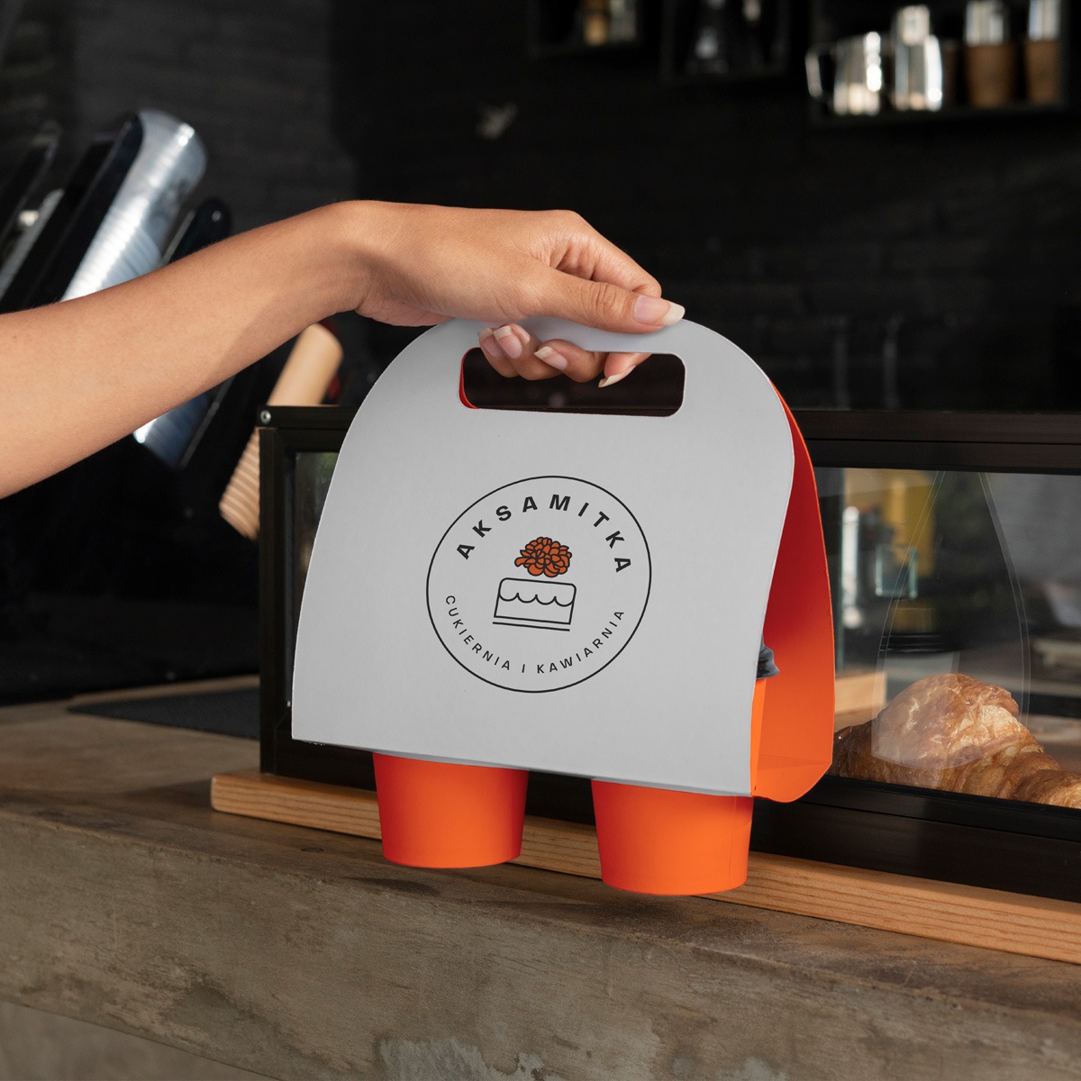 Dłoń trzyma papierowy uchwyt z logo 'Aksamitka' (cukiernia i kawiarnia) i dwoma pomarańczowymi kubkami na tle lady z croissantem w witrynie.