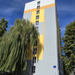 Wysoki blok mieszkalny po metamorfozie, z szaro-żółtą elewacją i białymi oknami, częściowo zasłonięty drzewem. Błękitne niebo w tle.