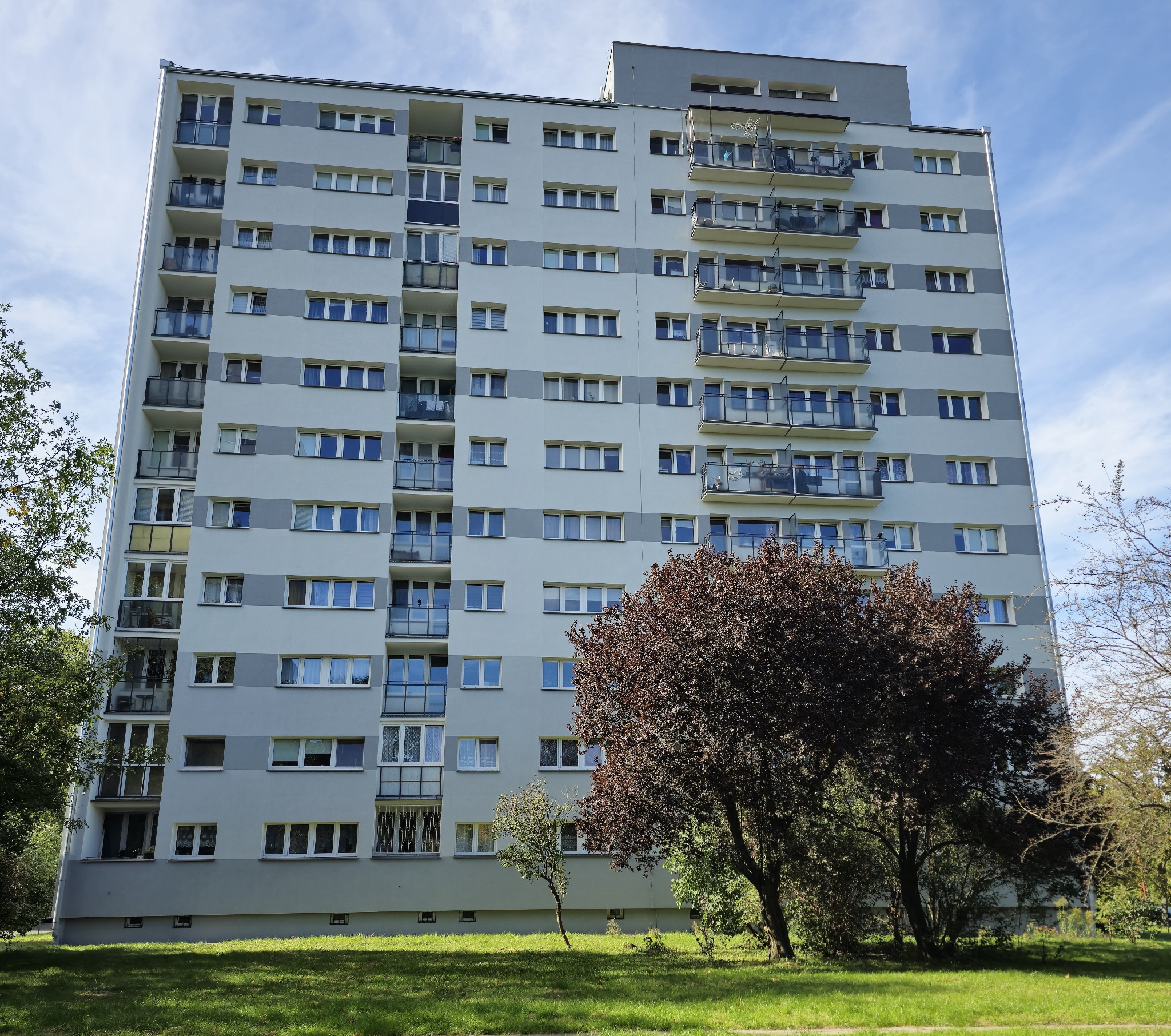 Wielopiętrowy blok mieszkalny po renowacji elewacji, z szarymi pasami i balkonami, otoczony zielenią. Widok z poziomu trawnika, błękitne niebo w tle.