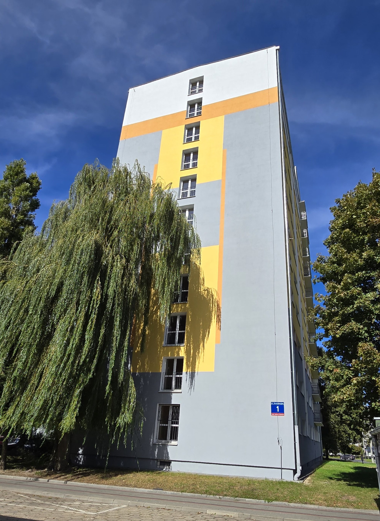 Wysoki blok mieszkalny po metamorfozie, z szaro-żółtą elewacją i białymi oknami, częściowo zasłonięty drzewem. Błękitne niebo w tle.