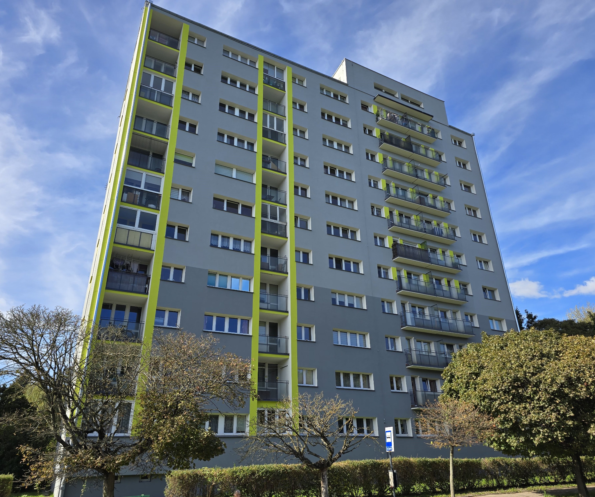 Wielopiętrowy budynek mieszkalny w szaro-zielonej kolorystyce, widziany z dołu pod kątem. Widoczne balkony i okna. Na pierwszym planie drzewa i krzewy.