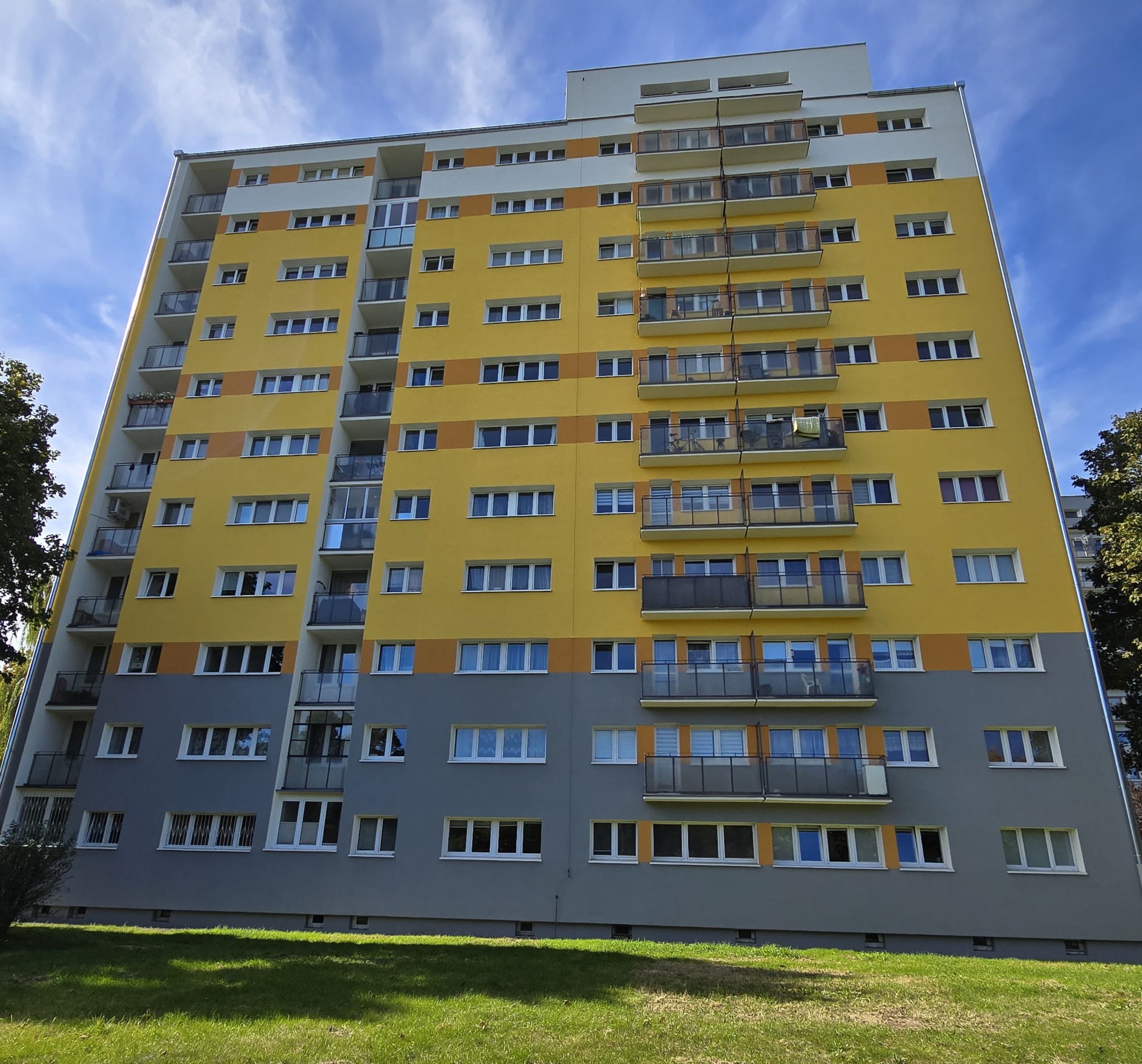 Wielopiętrowy budynek mieszkalny z balkonami, pomalowany w odcieniach żółci i szarości, z widoczną zielenią trawnika i drzew w tle, uchwycony w słoneczny dzień.