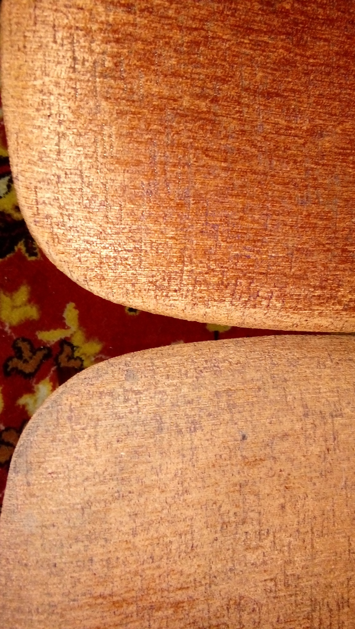 Fragment siedziska krzesła z pomarańczową tapicerką na tle wzorzystego dywanu. Widoczne ślady użytkowania na tkaninie.